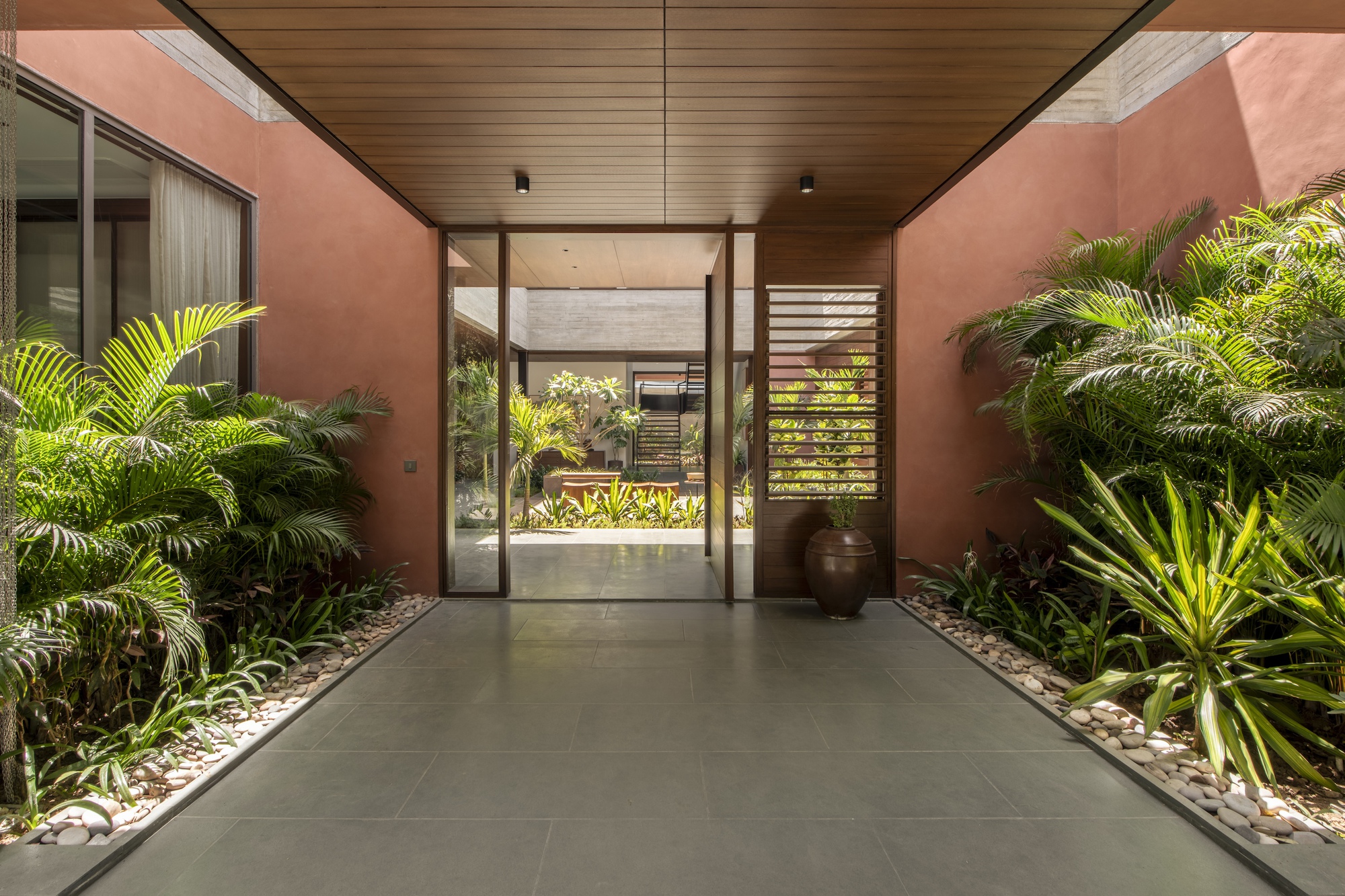 Entry passage with pebble-edged palm beds leading to a glass pivot door and timber ceiling, courtyard visible beyond