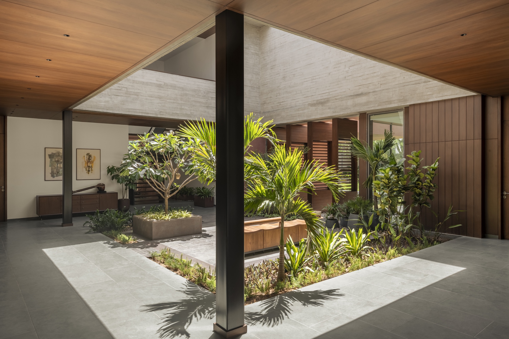 Black steel column in front of a courtyard garden, casting palm shadows across grey floor tiles beneath a timber soffit