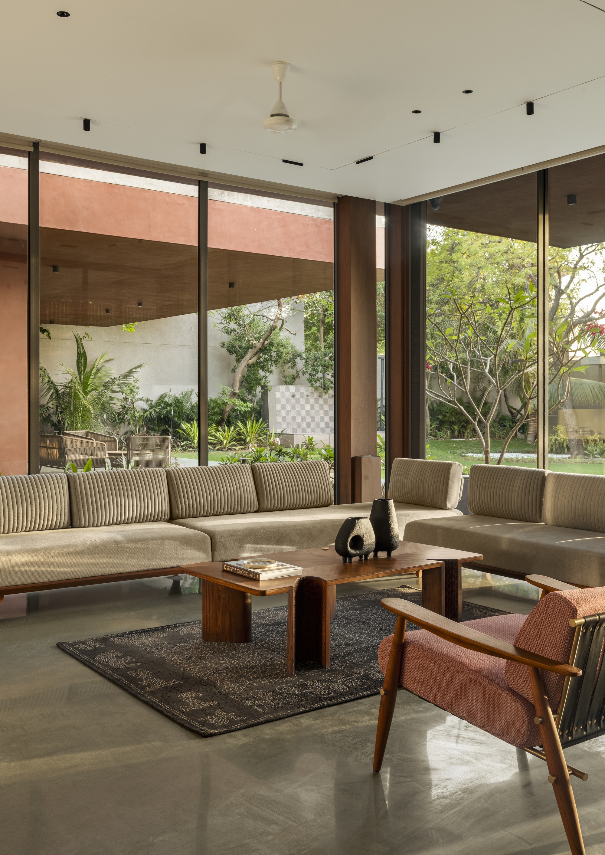 Living room with low striped sofas, a walnut coffee table and a pink armchair facing floor-to-ceiling glass and garden trees in warm light