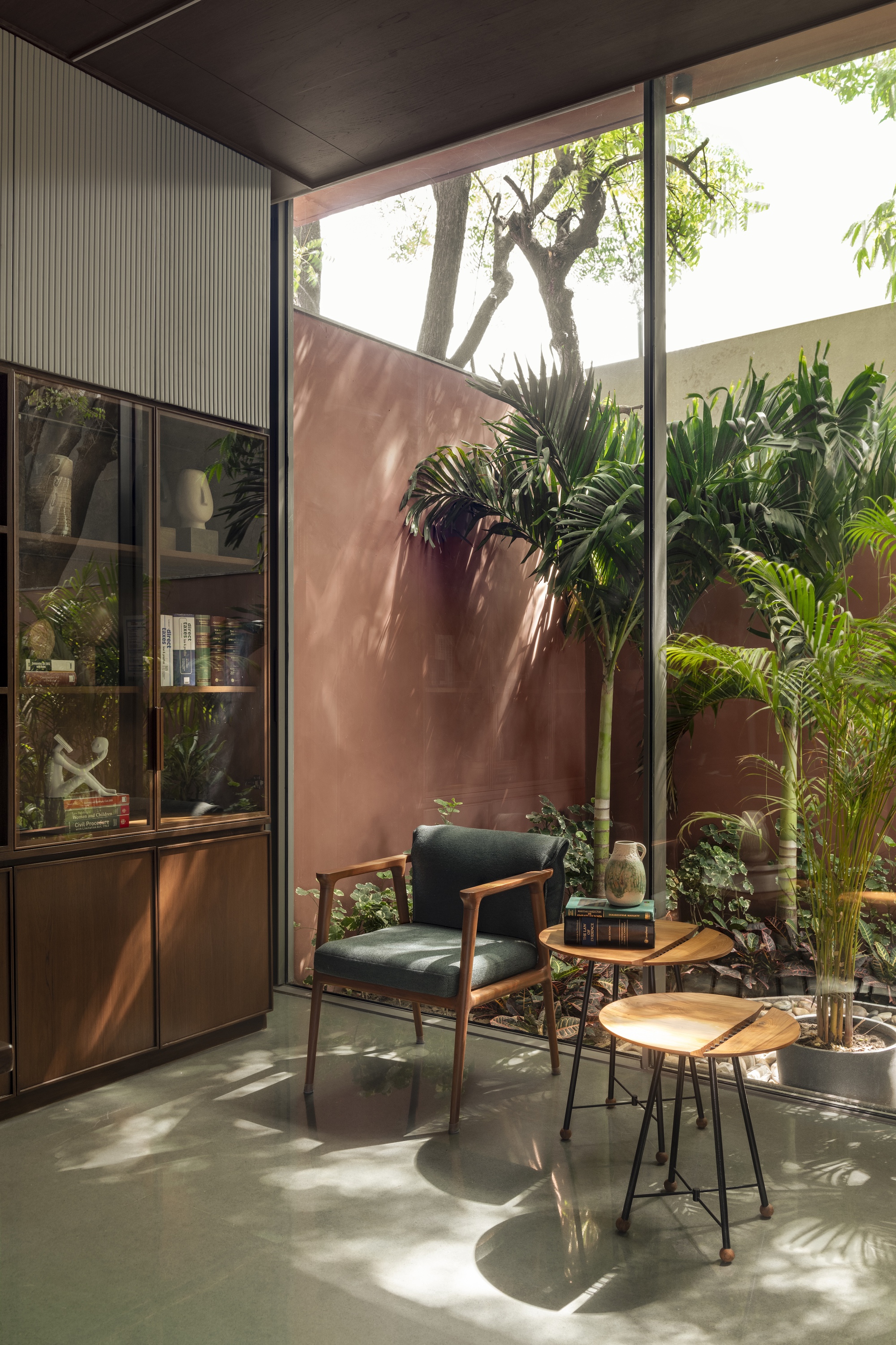 Reading corner with a green armchair and round side tables beside a glass wall, dappled sunlight falling on the polished floor