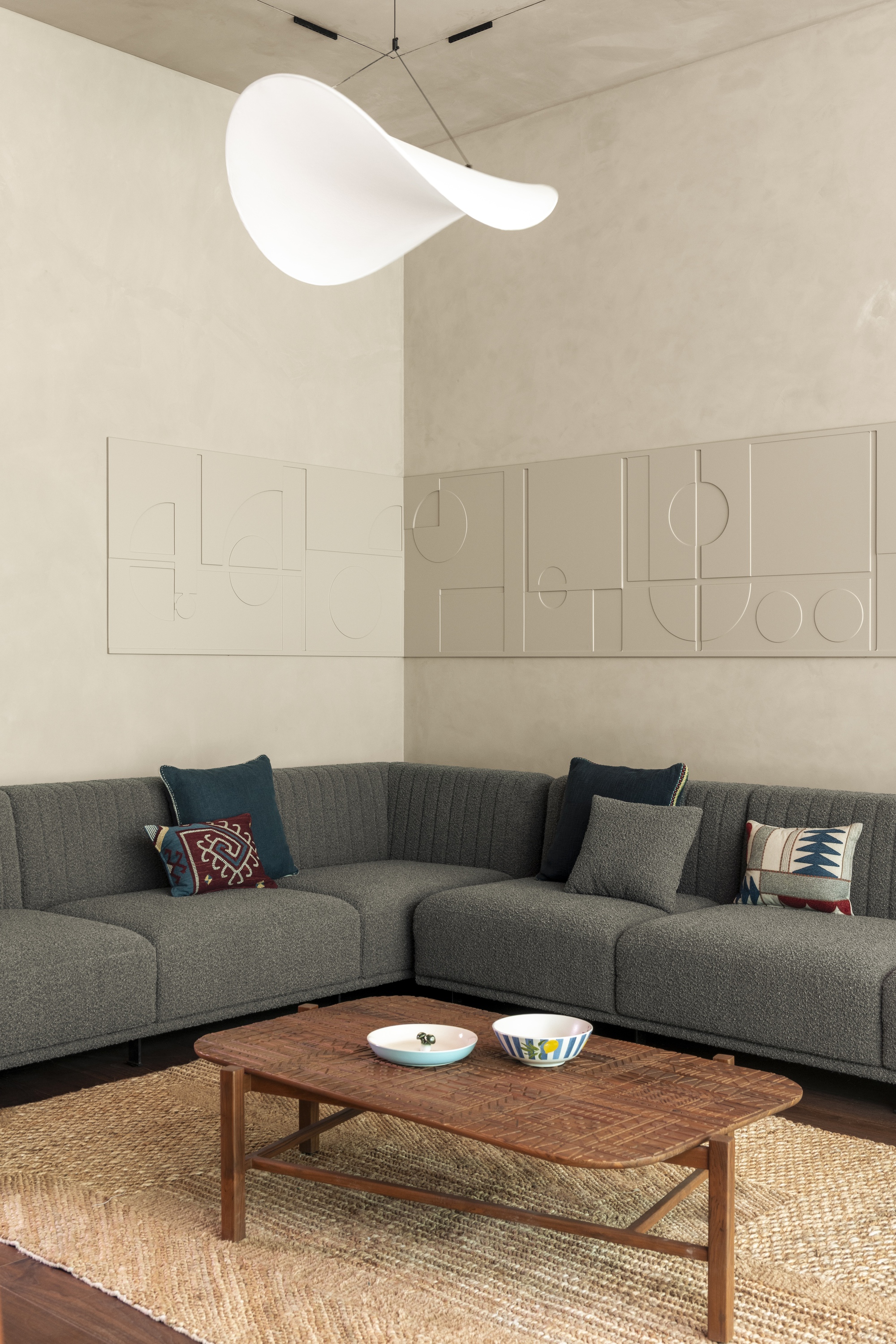 Corner of a grey bouclé sectional sofa beneath relief wall panels and a white sculptural pendant lamp, with a jute rug below