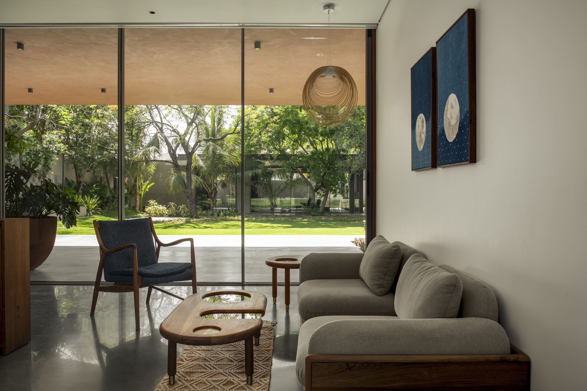 Seating area with a grey sofa and blue armchair facing a glass wall that opens onto a lawn and mature trees