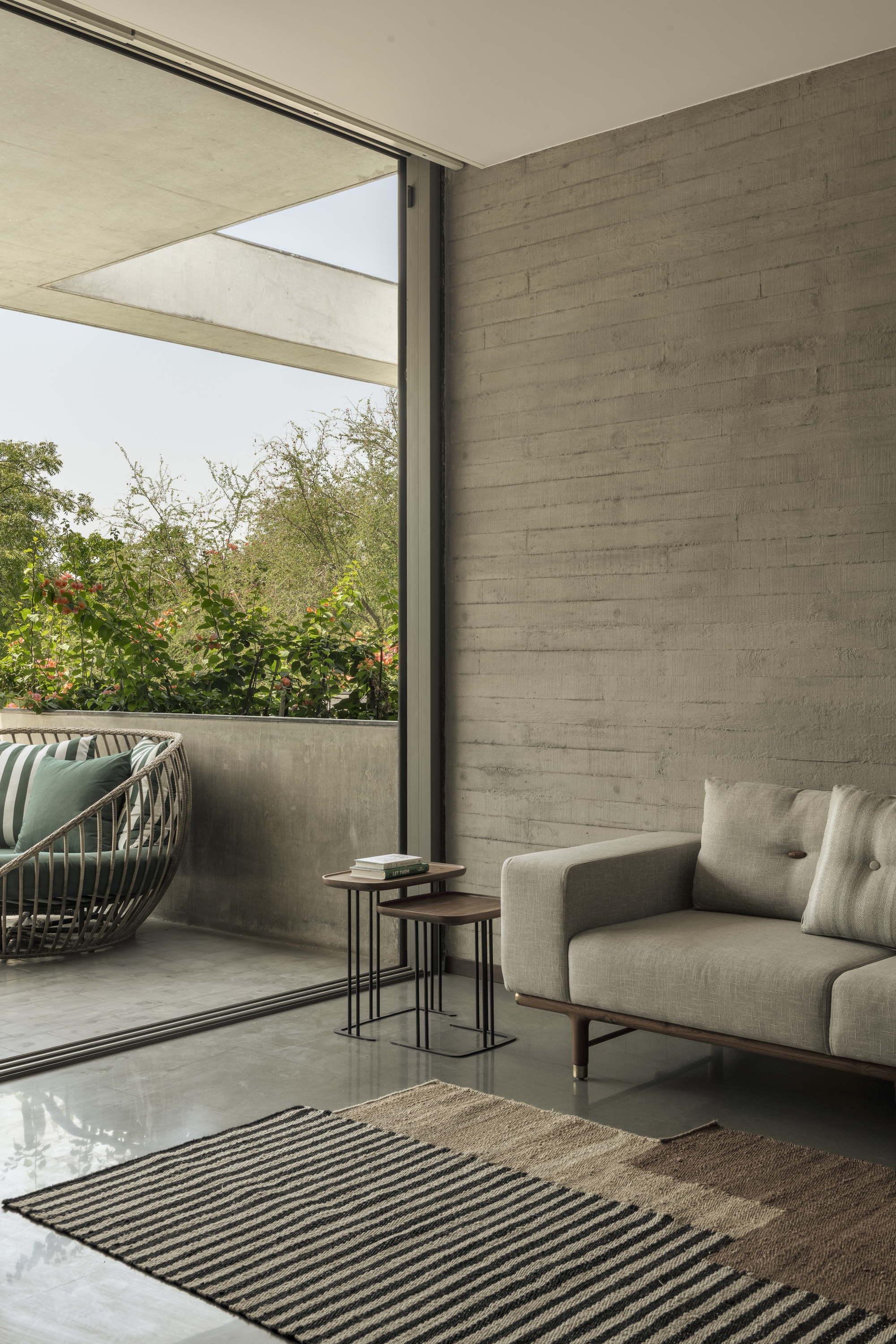 Board-formed concrete wall beside a sliding glass door, with a woven balcony chair and bougainvillea outside in soft daylight