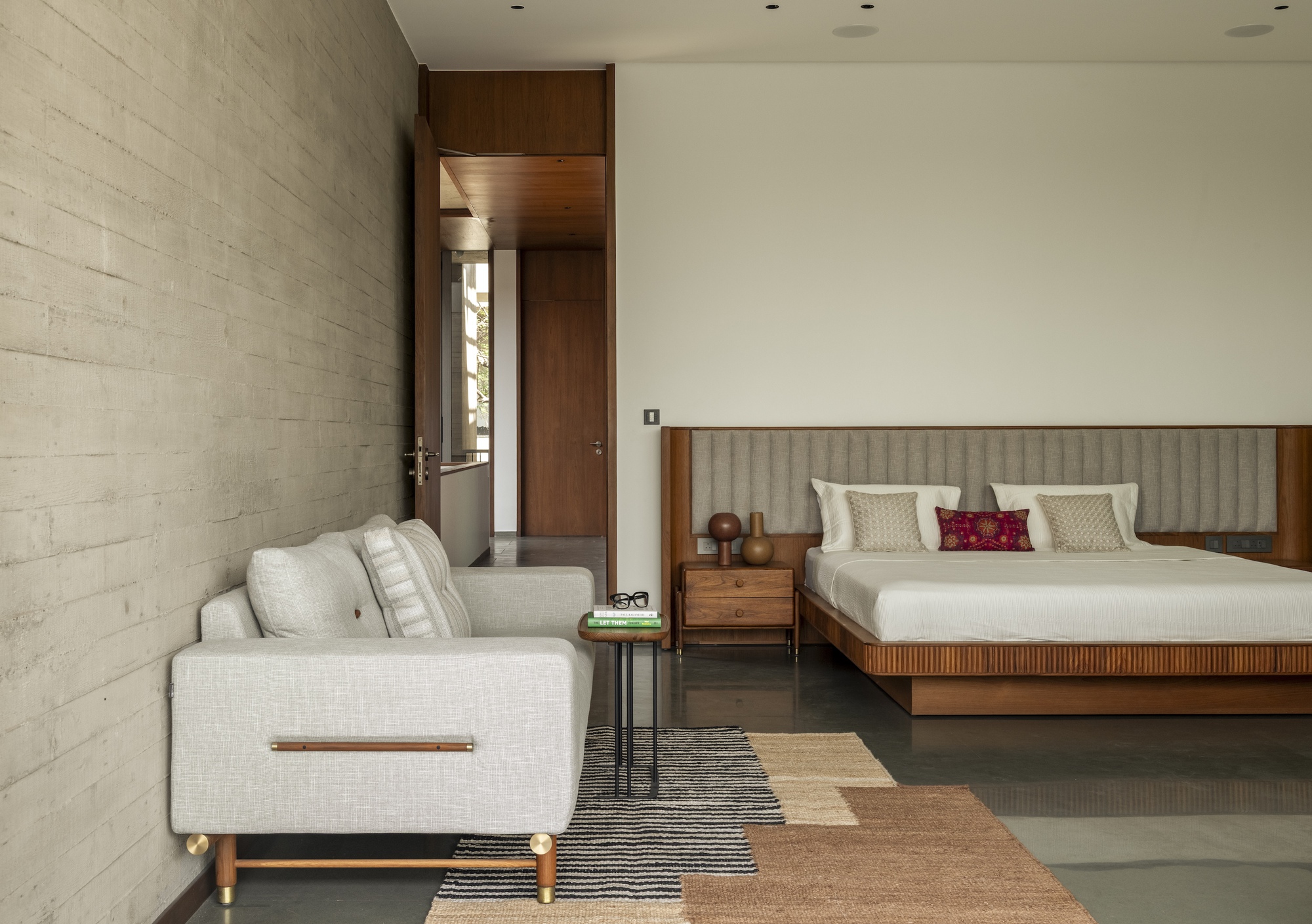 Bedroom with a timber platform bed, padded headboard and board-formed concrete wall, with a pale sofa on striped rugs