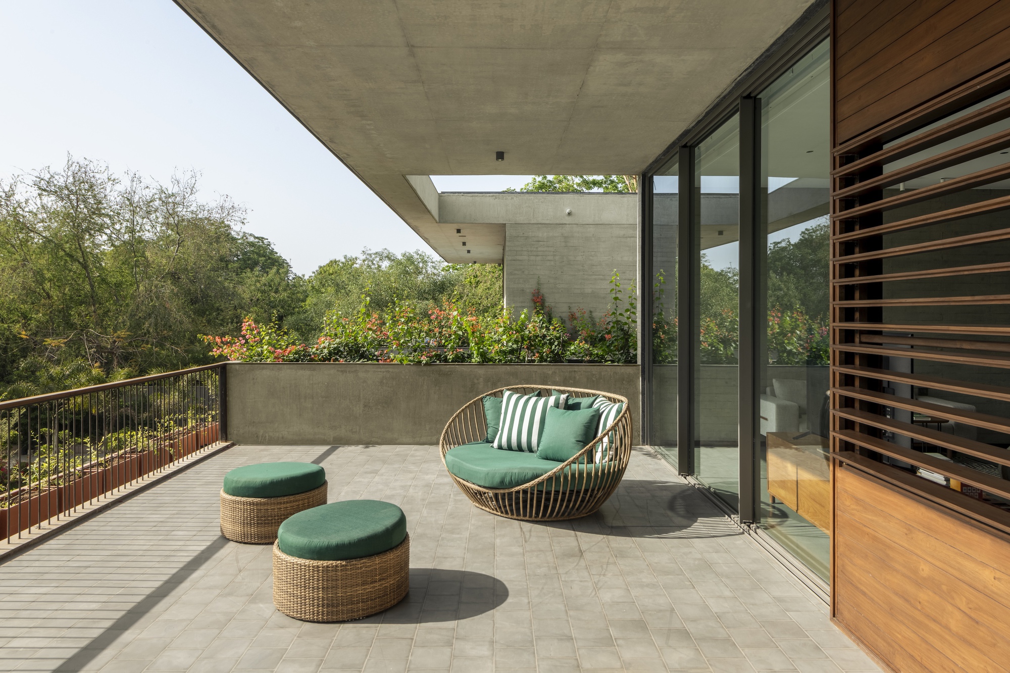 Concrete-roofed terrace with a rattan daybed, two green round ottomans and a planted parapet against a backdrop of trees