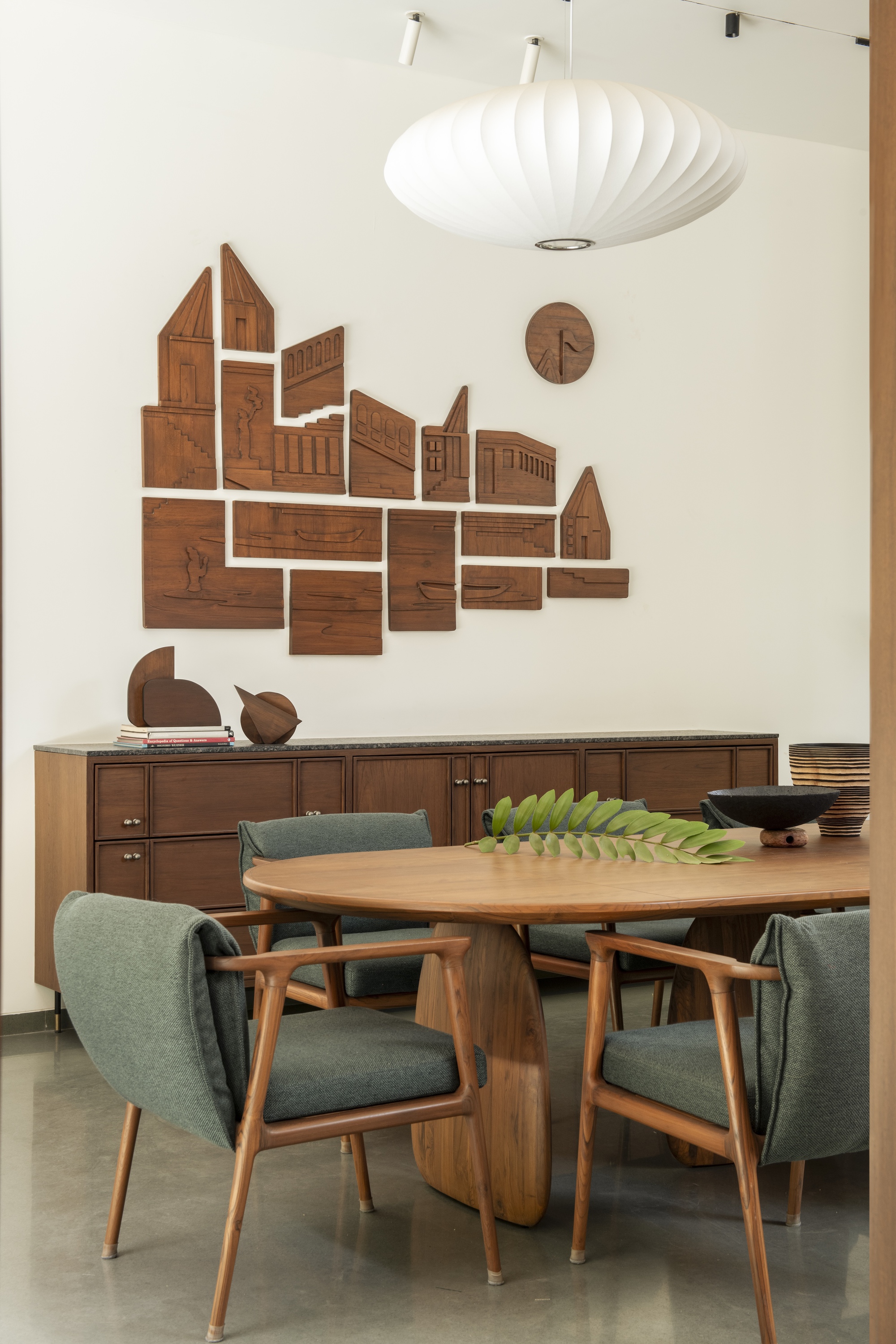 Dining area with an oval timber table, green upholstered chairs and a carved wooden relief of buildings above a sideboard