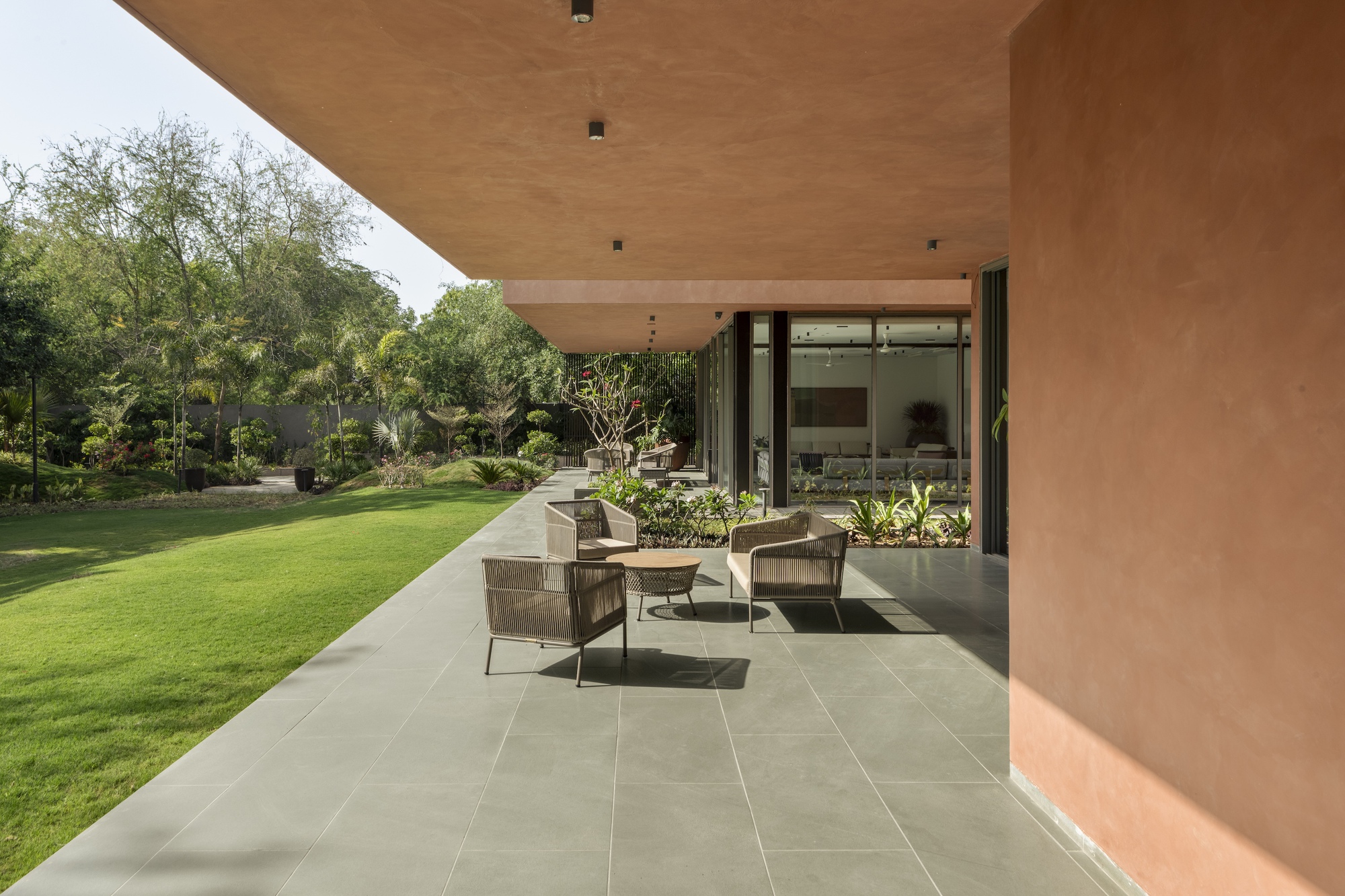 Covered terrace with terracotta-toned plastered walls and cantilevered roof, wicker chairs on grey stone paving facing a lawn in afternoon sun