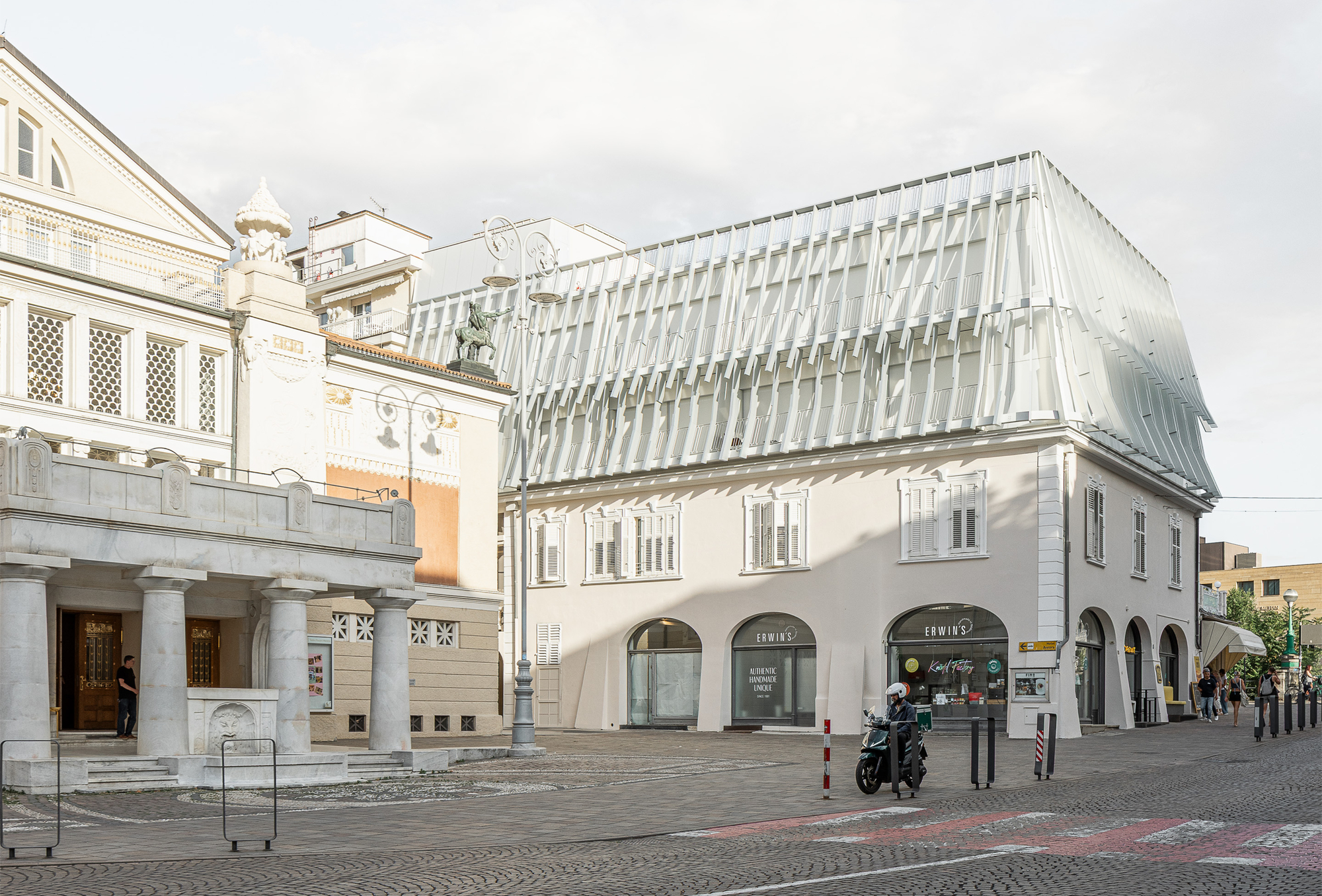 Corner view of the white plastered building with a pleated, translucent upper roof volume beside a columned portico, a scooter parked below on cobbles