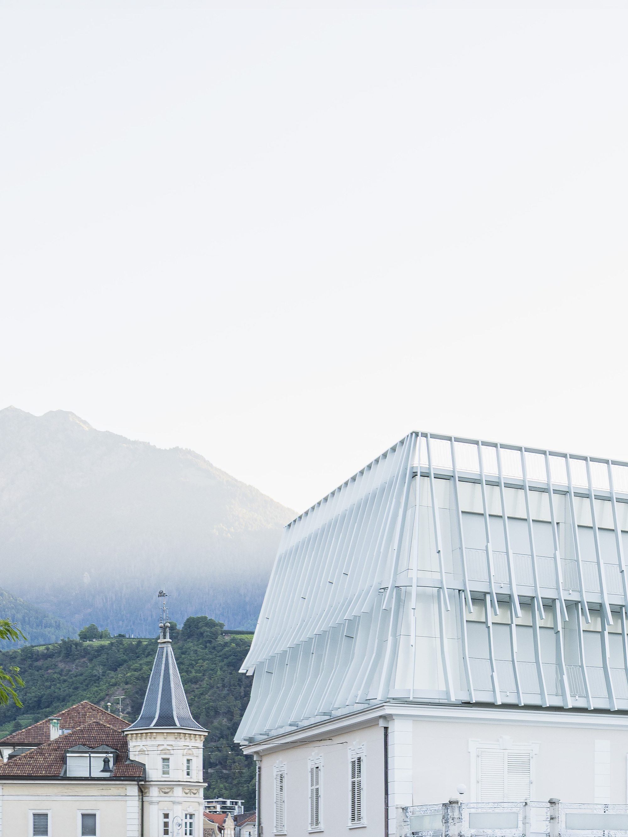 Pleated white metal roof volume rising above neighboring rooftops, with a misty forested mountain slope behind in soft morning haze