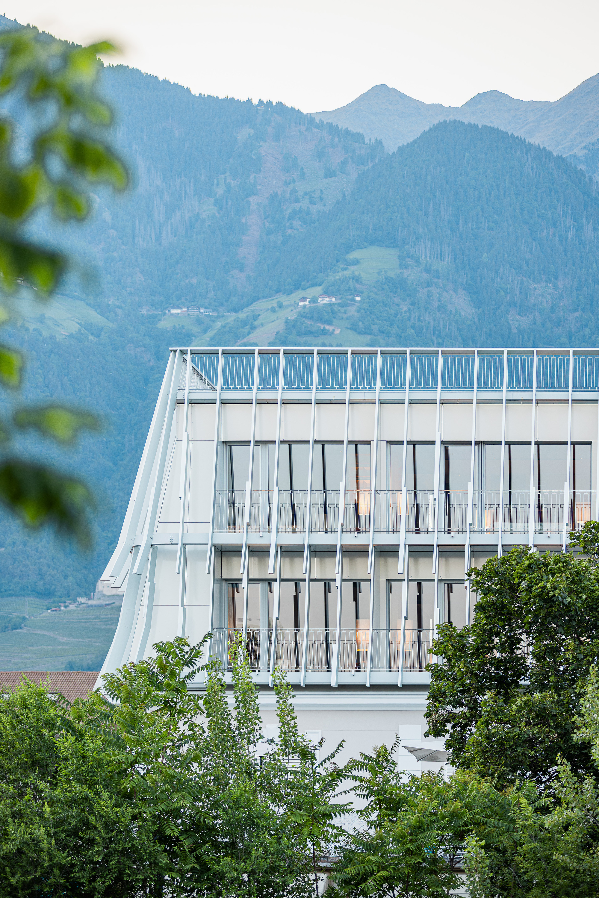 Upper floors with slender vertical fins and balcony railings, framed by leafy branches against a blue forested mountainside