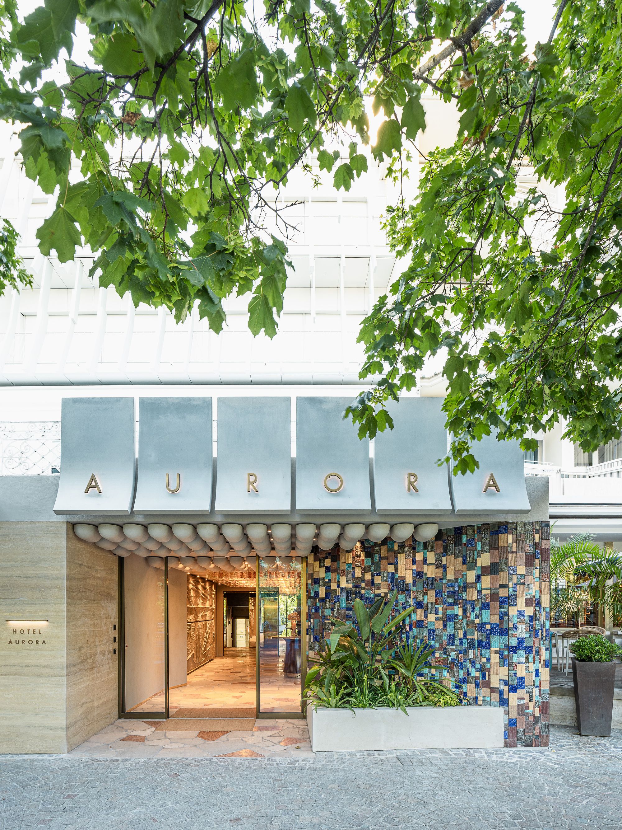 Hotel entrance with curved metal canopy panels, gold lettering and a mosaic tile wall, shaded by overhanging green leaves