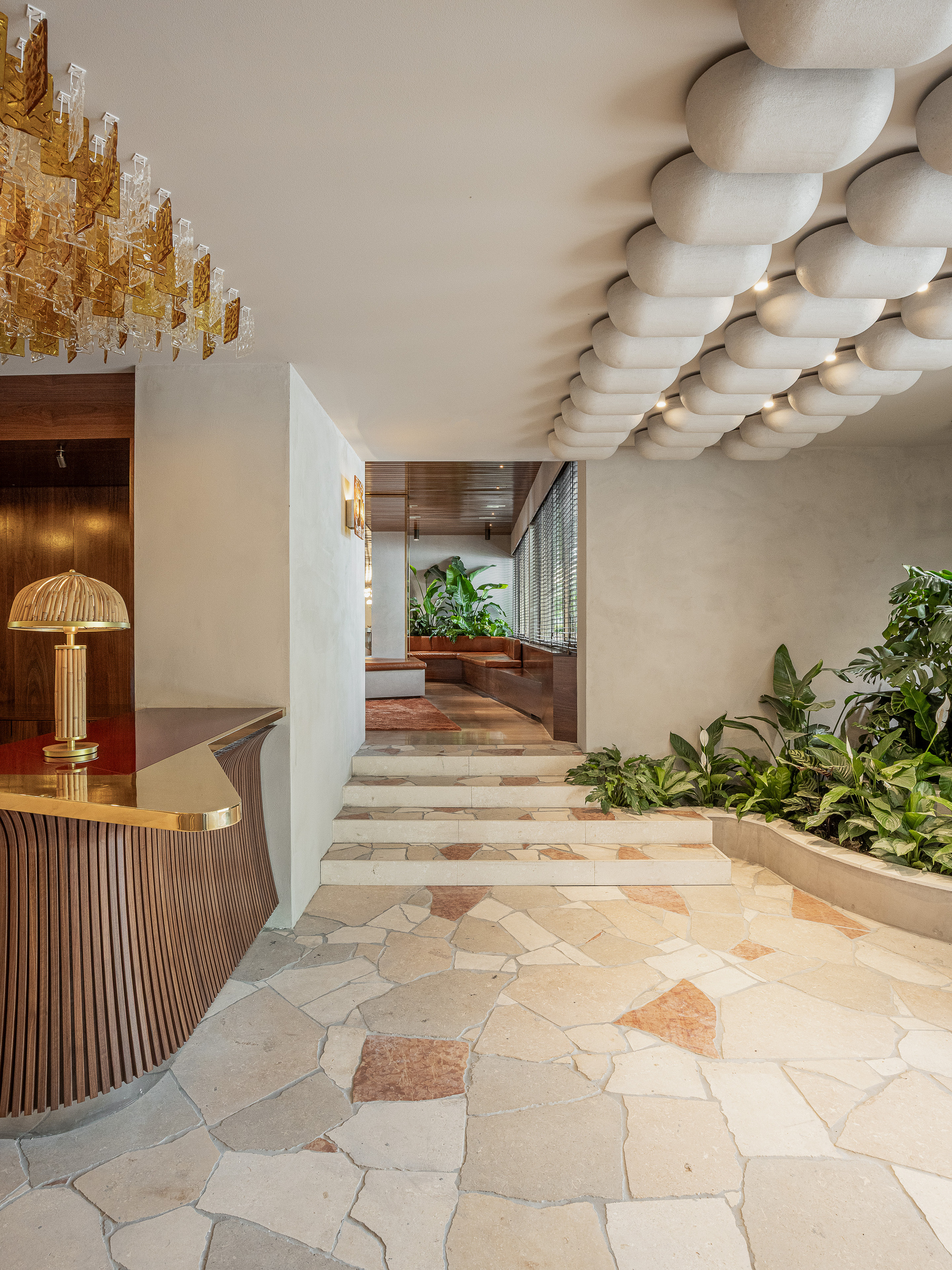 Lobby with a brass-topped curved timber reception desk, pillowy ceiling pods, irregular stone paving and a planted bed beside low steps