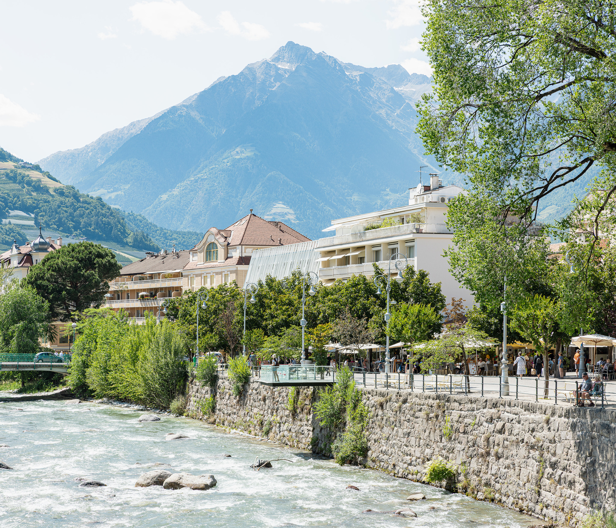 River running past a stone embankment wall, with a promenade of café umbrellas, trees and a snow-dusted peak behind in summer sun