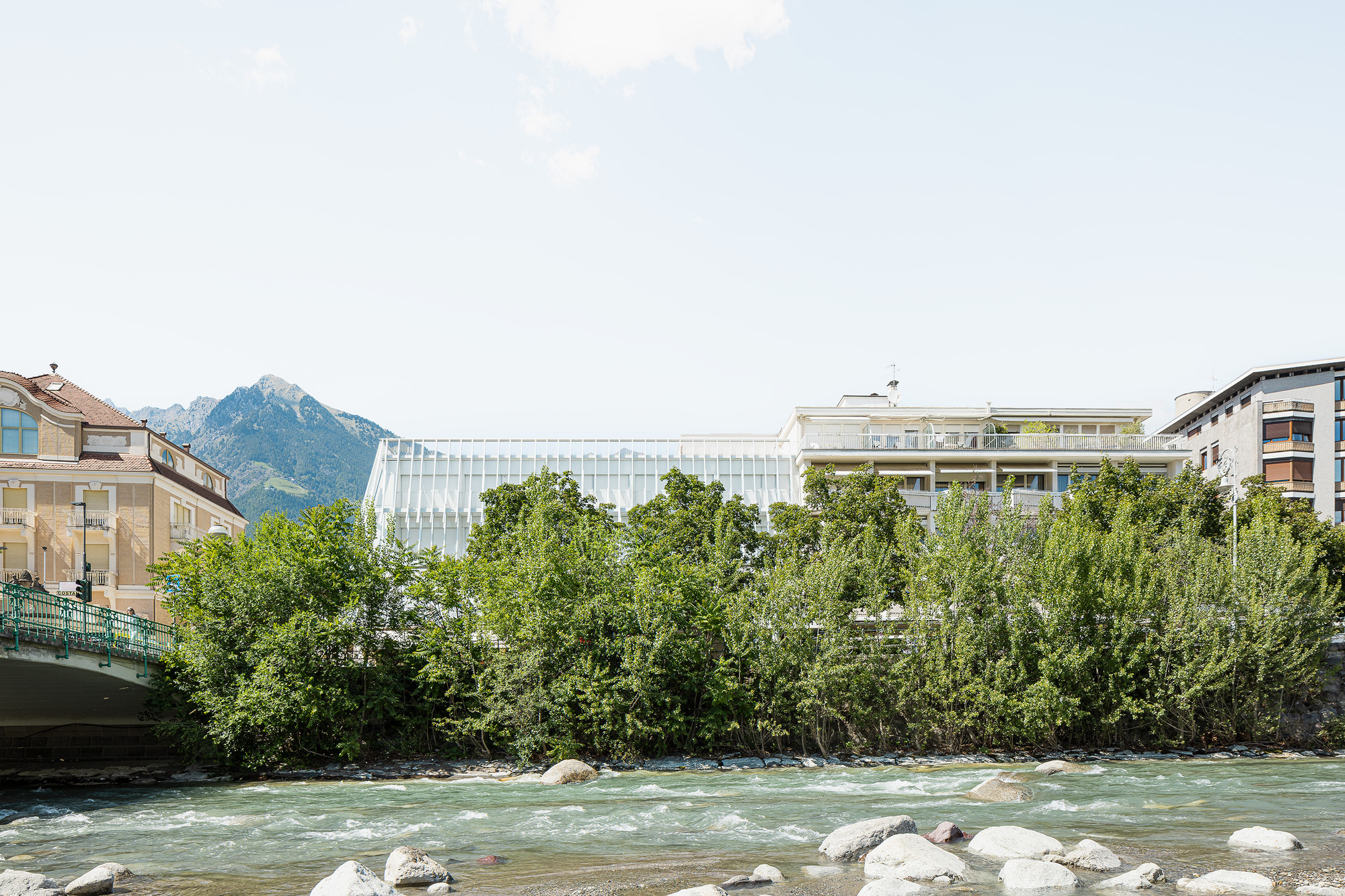 White glazed rooftop volume rising behind a row of riverside trees, with rapids and rounded boulders in the foreground under a clear sky