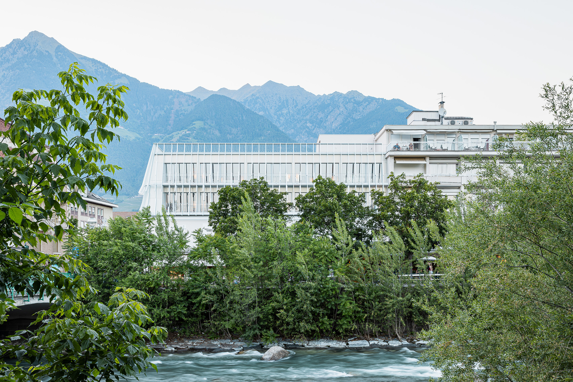 Glass-fronted upper floors with fine vertical fins seen across a rushing river, framed by leafy branches and mountains at dusk