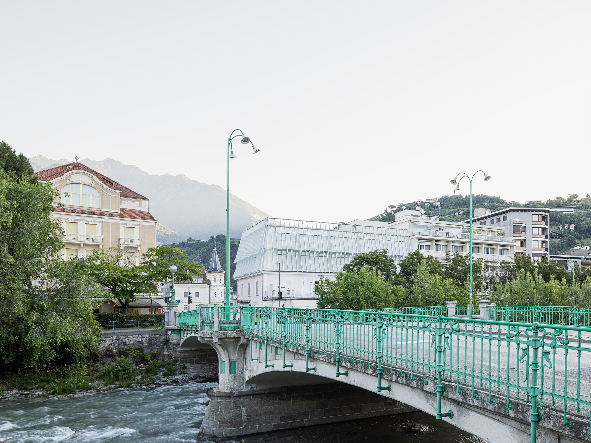 Green cast-iron bridge railing leading toward a pale ribbed glass roof, with a mountain ridge behind in soft evening haze