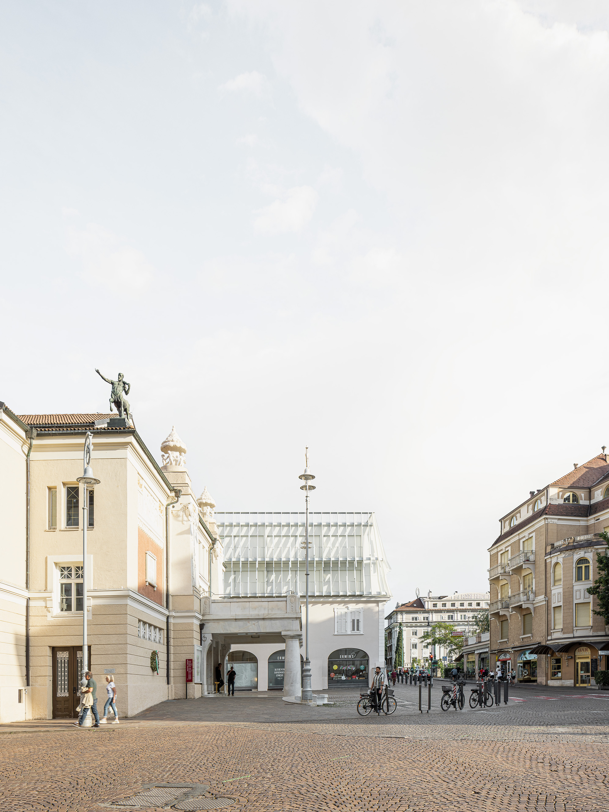 Cobbled square with a glazed pavilion volume set between historic facades, cyclists and pedestrians crossing under a pale cloudy sky