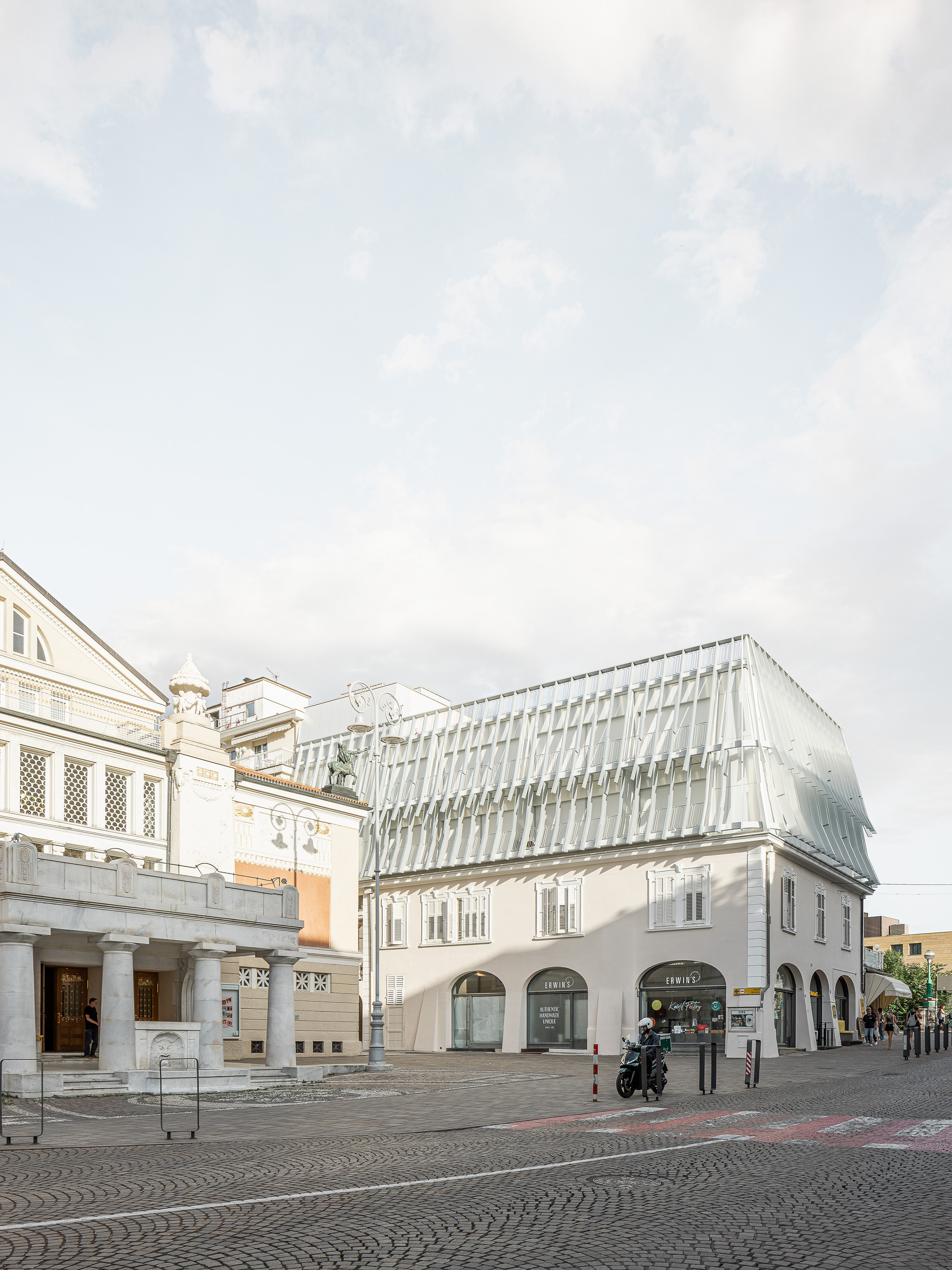 Corner building with arched ground-floor shopfronts and a folded, finned translucent roof, a scooter parked on the cobbles in afternoon light