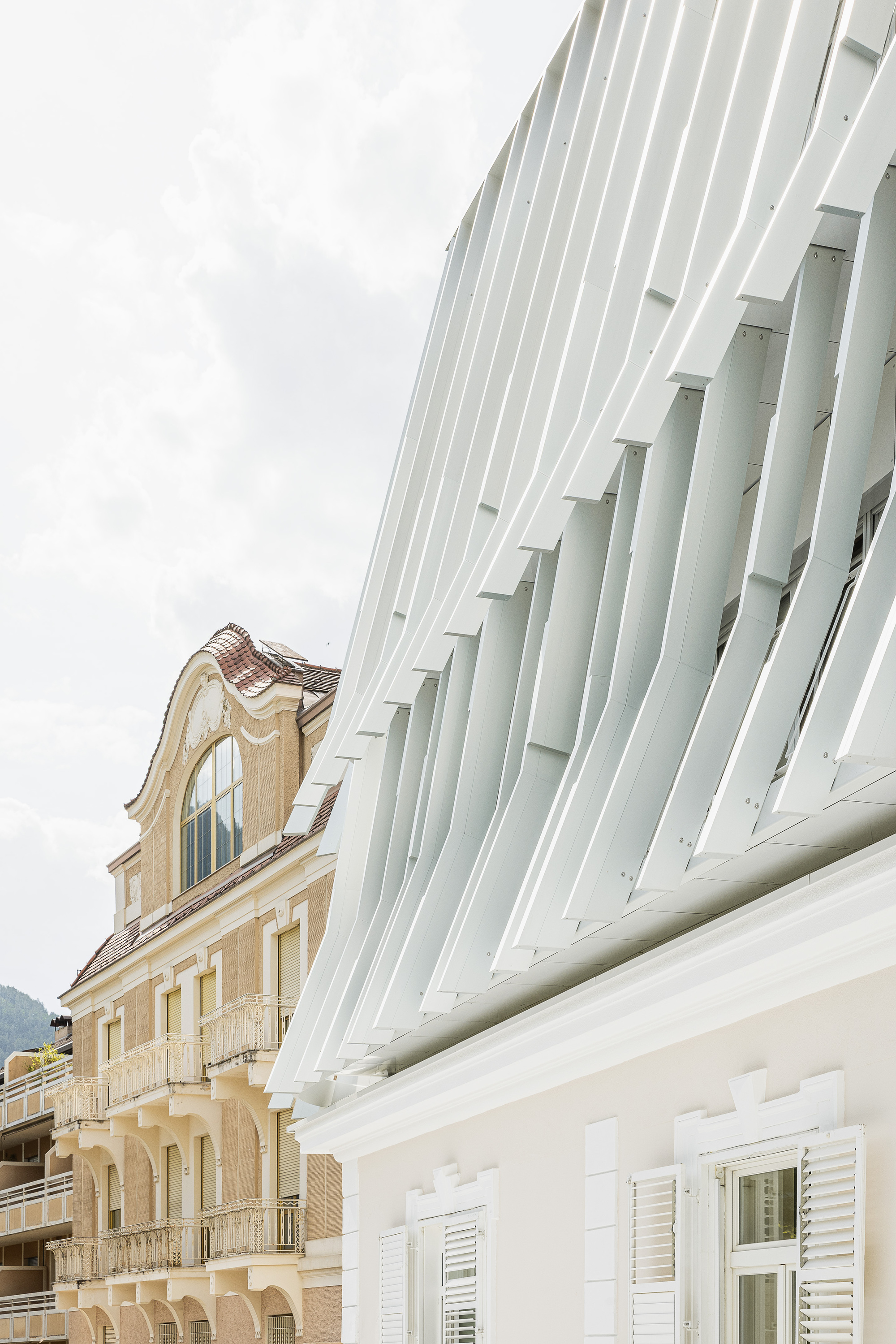Close-up of overlapping white vertical fins curving over a plastered cornice with shuttered windows, an ornate ochre facade beside it under cloud