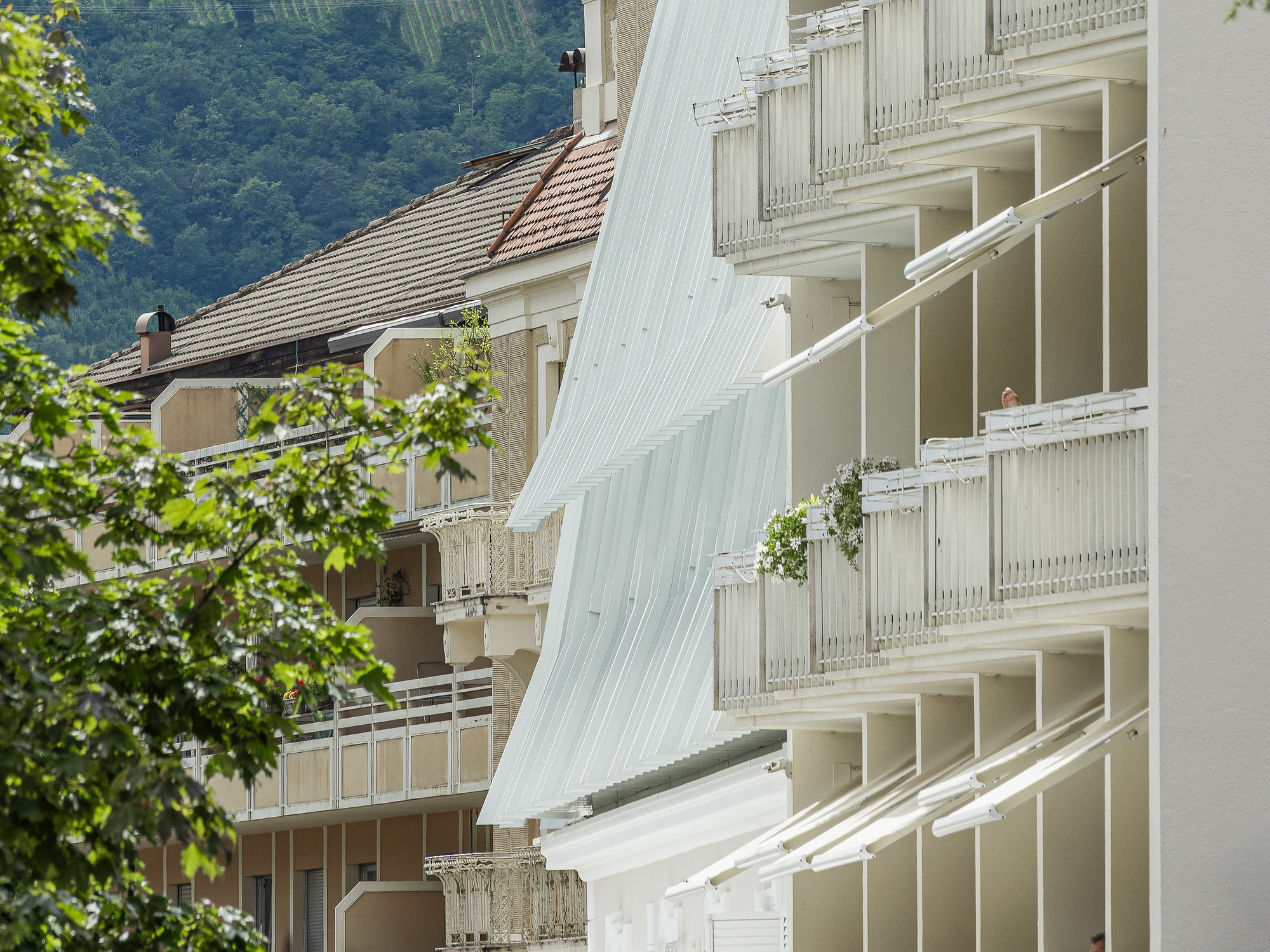 Pleated white metal screen beside balconies with white railings, set against tiled rooftops and a wooded hillside in summer leaf