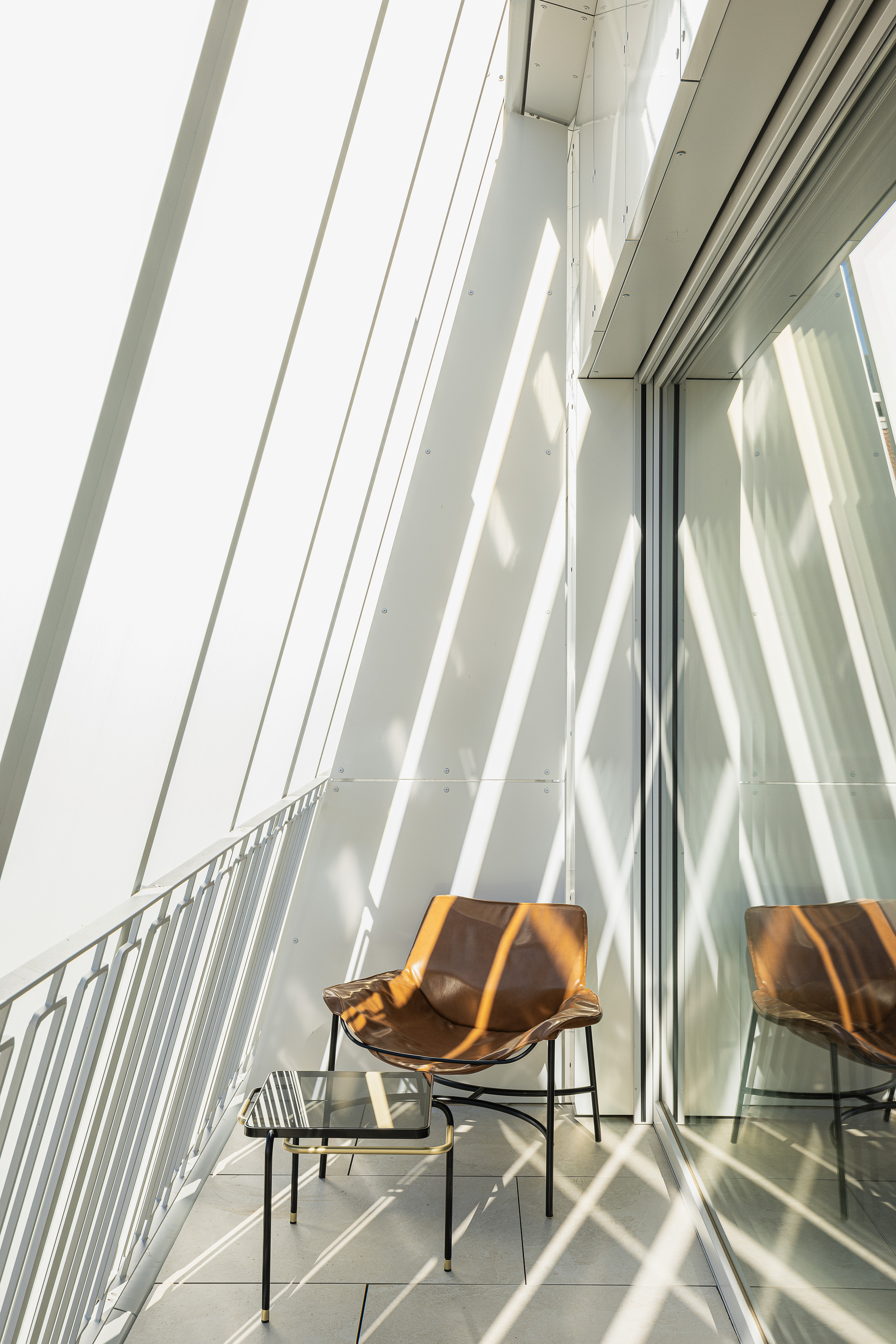 Narrow balcony with a tan leather chair and glass side table, striped by sunlight through slanted white fins
