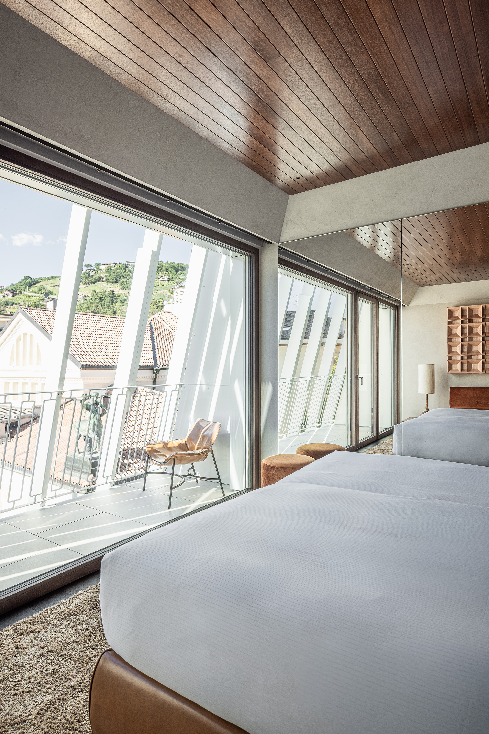Hotel room with timber ceiling and floor-to-ceiling glazing opening onto a balcony, with a hillside of vineyards under blue sky