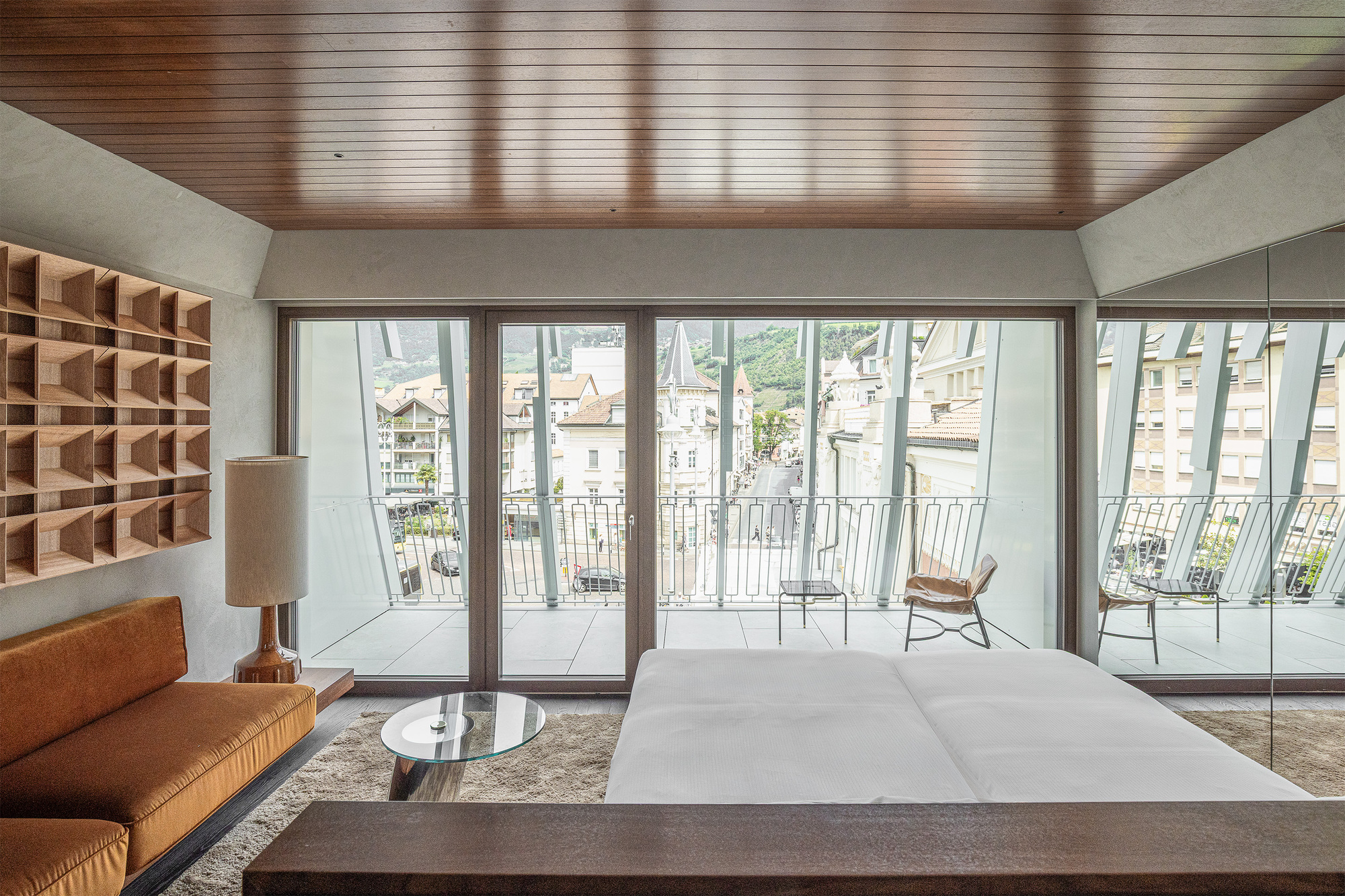 Bedroom facing a glass balcony door, with an ochre sofa, wall shelving and a street lined with pitched roofs beyond
