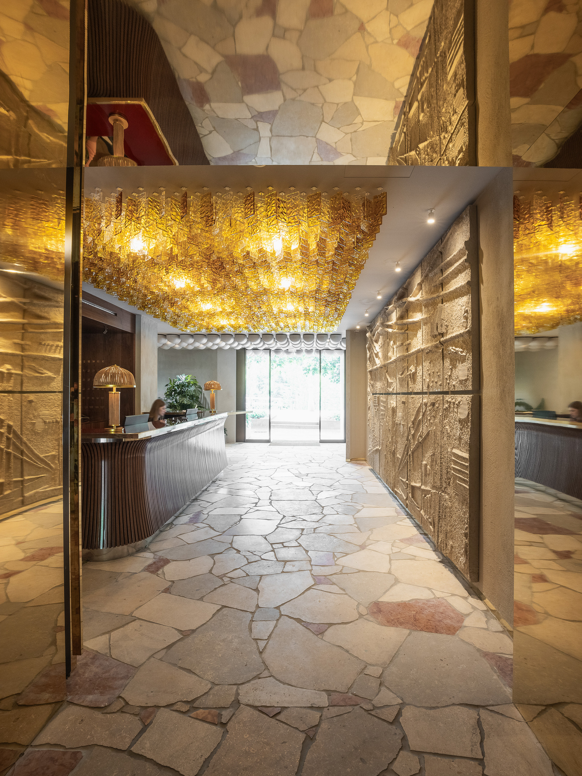 Lobby with irregular flagstone floor, amber glass ceiling cluster, curved reception desk and relief panels on the wall, daylight at the far entrance