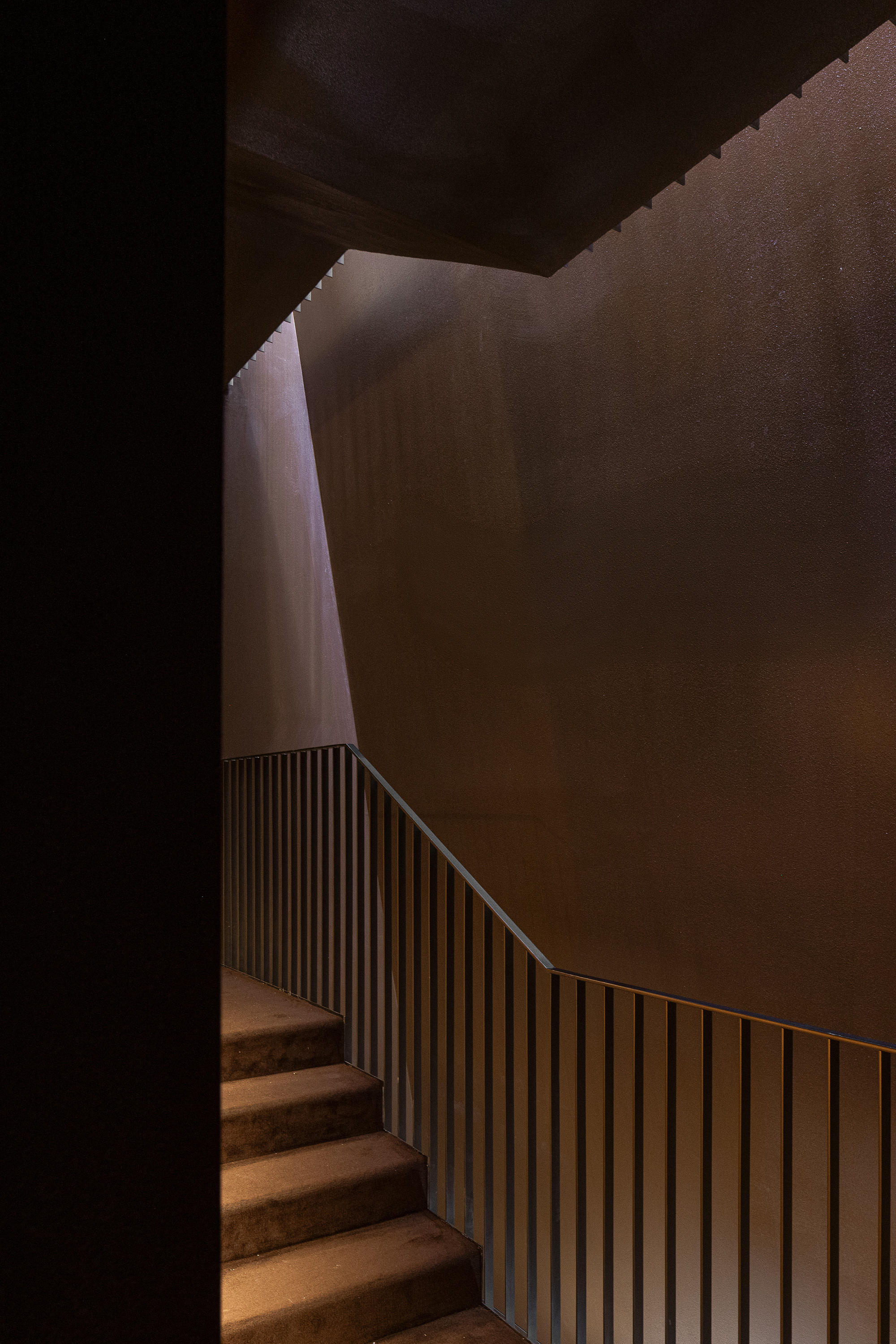 Stair flight with carpeted treads and slim vertical metal balustrade beside angled weathering-steel walls, lit by a thin shaft of daylight from above