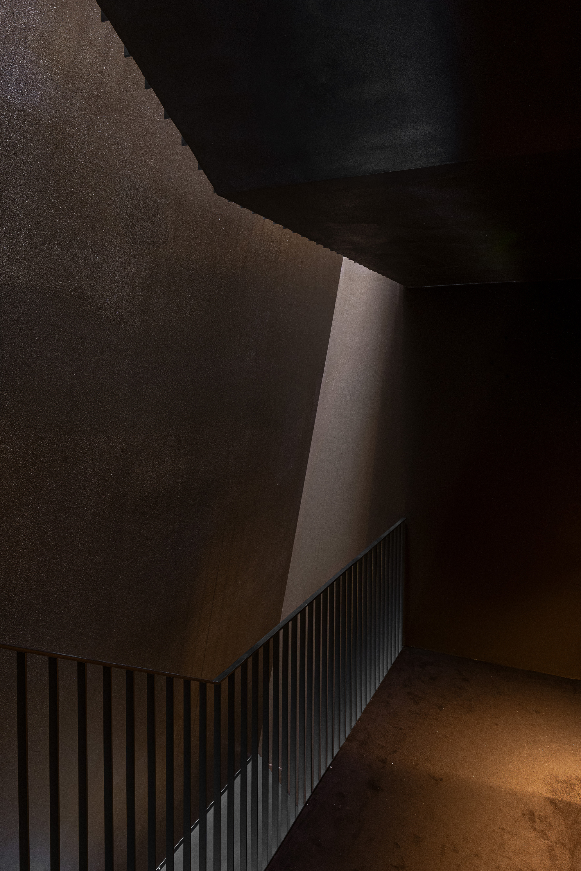 Landing with a vertical-bar railing below a slanted dark wall, where a sliver of daylight falls on the textured plaster