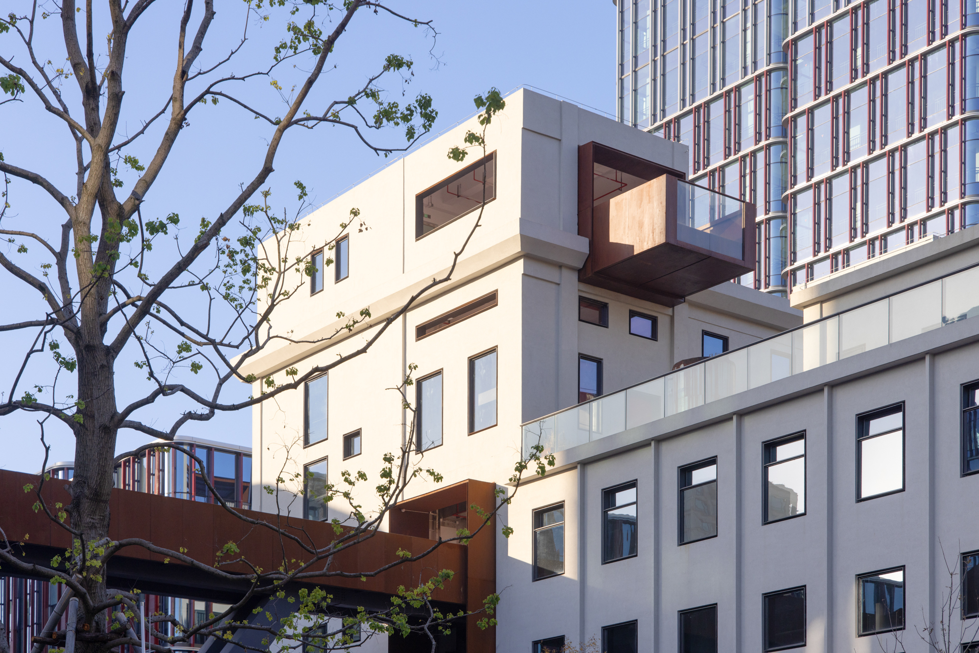 Cream-rendered stacked volumes with a cantilevered rust-colored steel box and glass balustrade, framed by bare branches against a clear blue sky