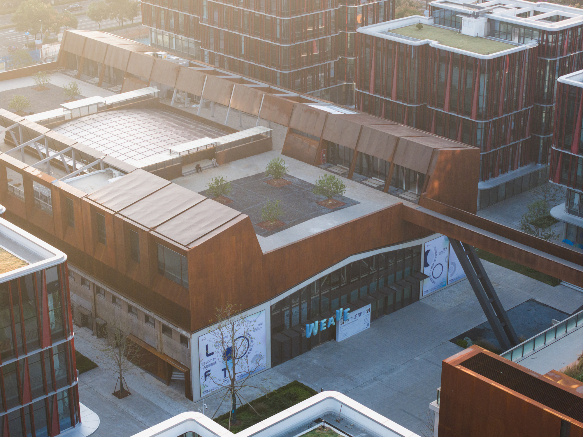 Aerial view of the weathering steel roof volumes, rooftop planting beds and a diagonal-braced bridge beside red-finned towers