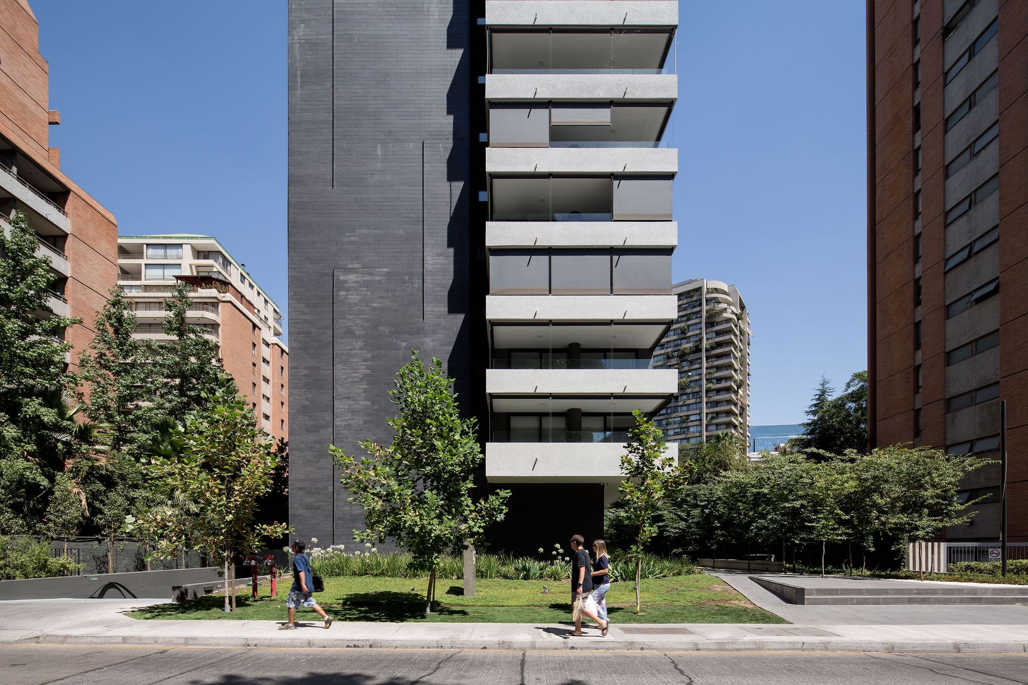 Street view of a dark slate-clad tower with stacked concrete balcony slabs, a lawn with young trees and pedestrians in midday sun