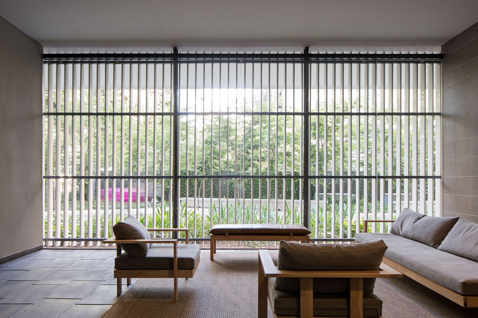 Lounge with wood-framed armchairs and a sofa facing a floor-to-ceiling vertical louver screen that filters green garden light