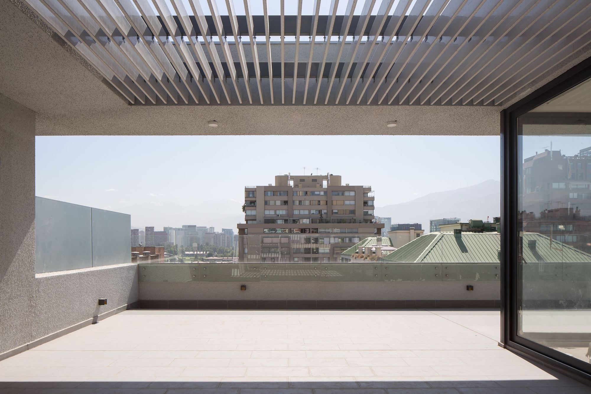 Rooftop terrace under a slatted pergola with a glass balustrade, framing neighboring towers and distant mountains in hazy sun
