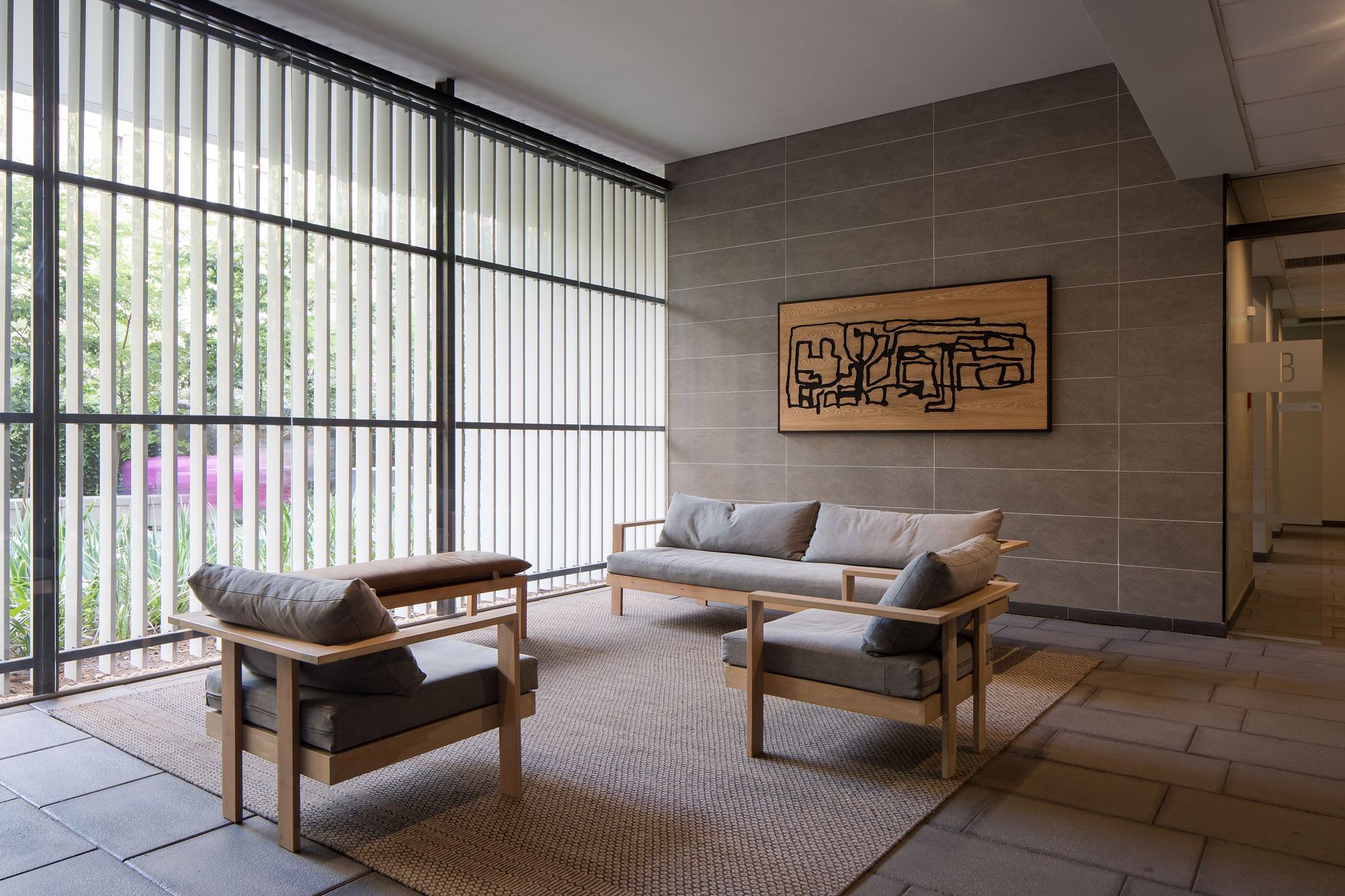 Lobby seating area with wood armchairs and sofa on a woven rug, a framed line artwork on a stone-tiled wall