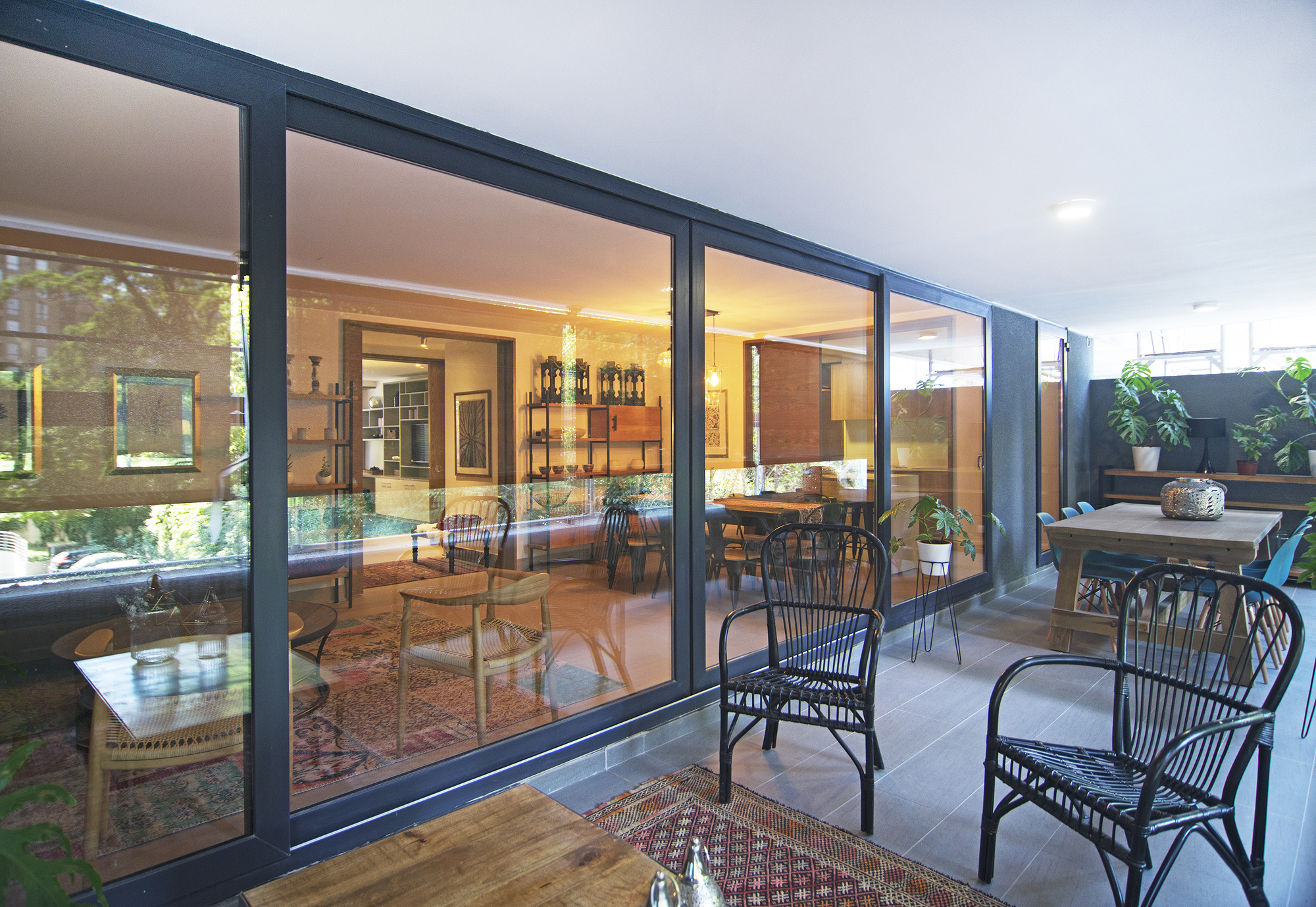 Covered terrace with black wicker chairs and a wooden table beside floor-to-ceiling sliding glass doors, warm lit living room beyond