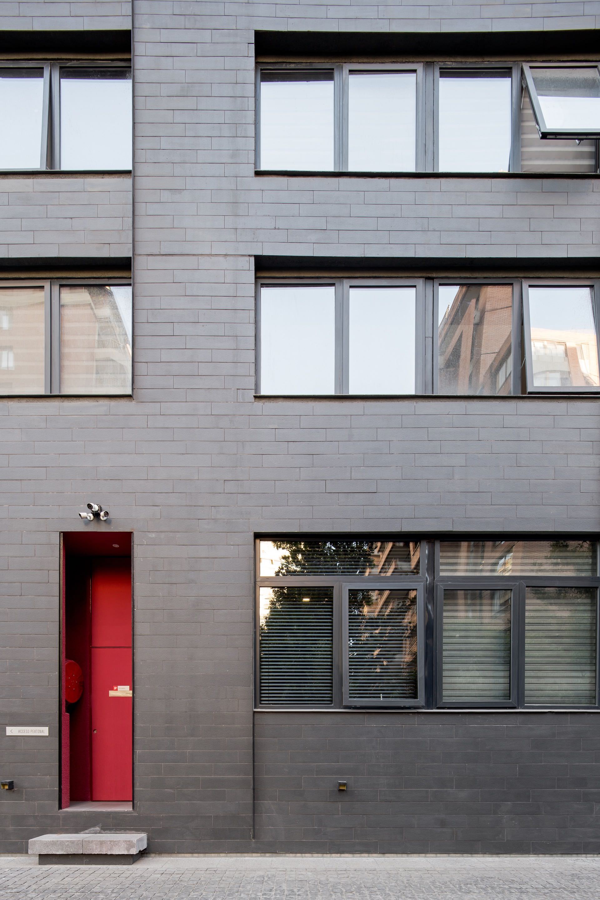 Dark grey brick facade with a narrow recessed red door, gridded windows and a stone step on a paved street