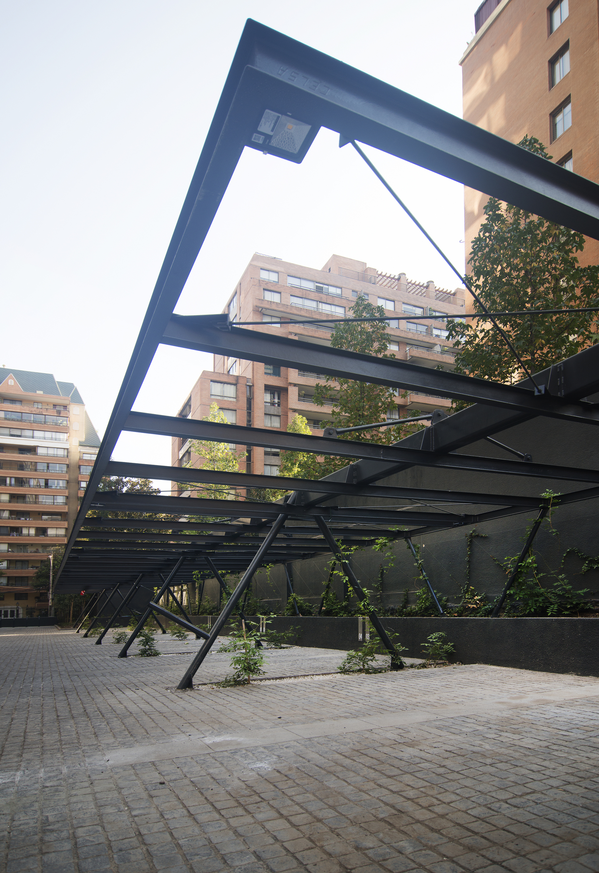 Low angle of a black steel pergola frame with diagonal struts over cobblestone paving, ivy-covered wall and brick towers behind in hazy daylight
