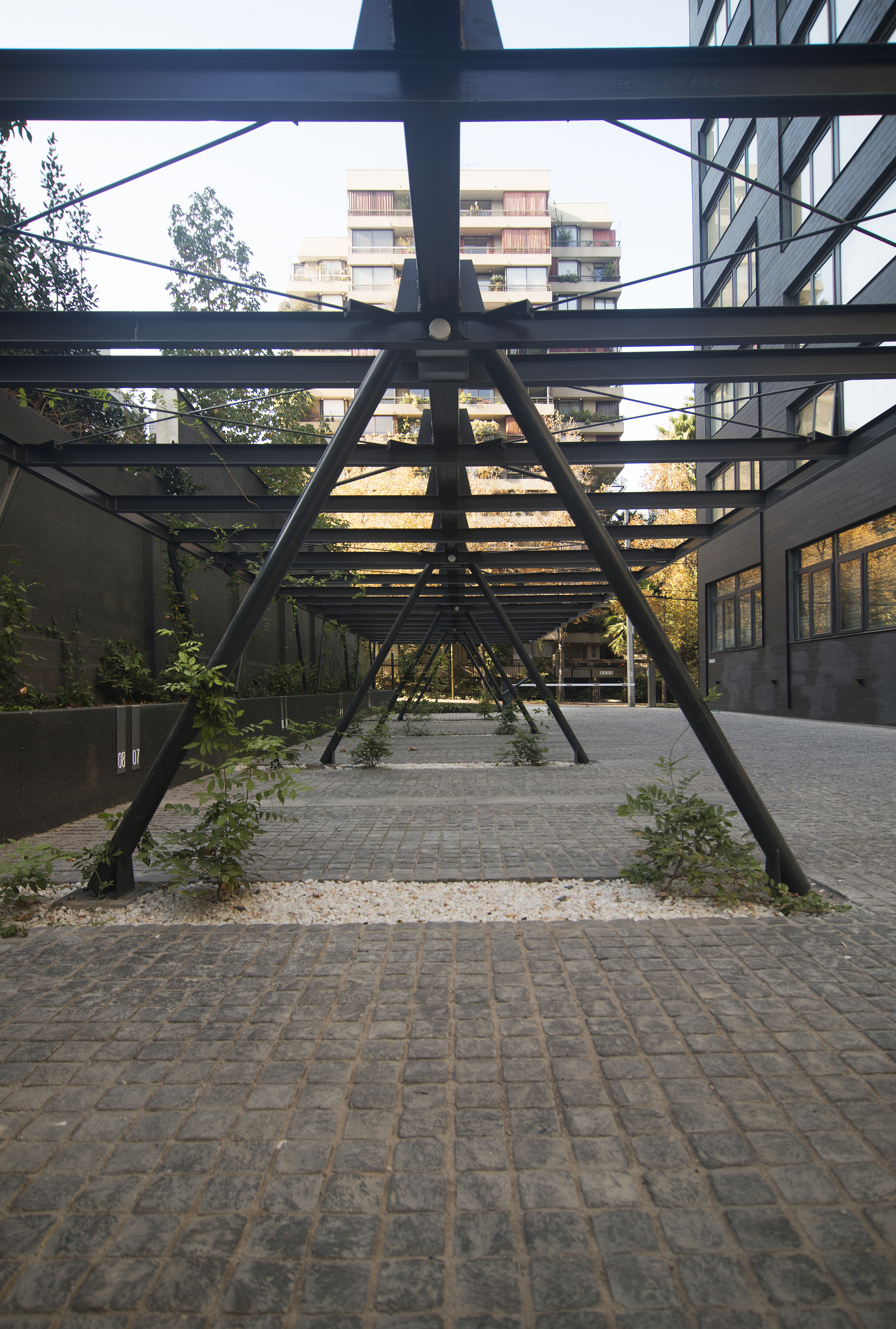 Looking down the steel canopy's repeating A-frame struts, with saplings in gravel strips between cobblestone paving and a dark facade at right