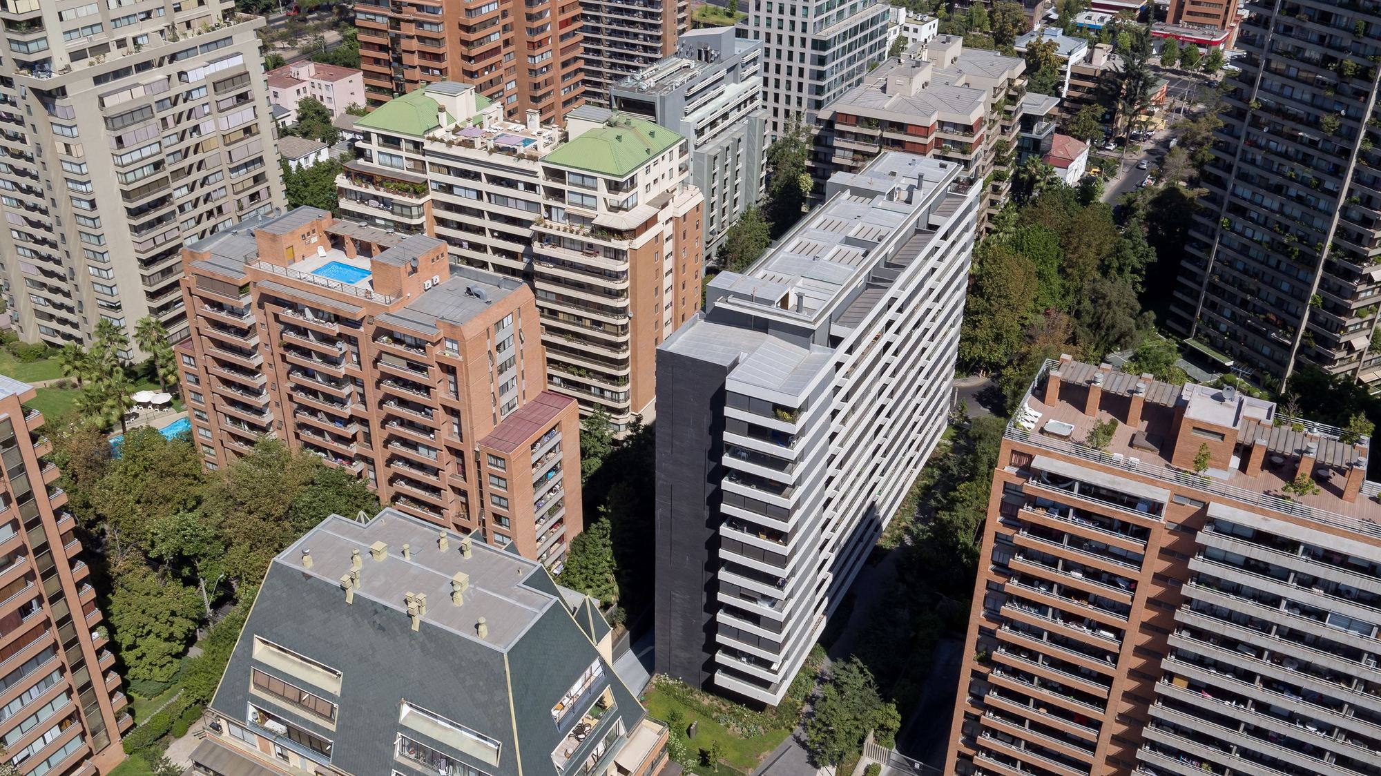 Aerial view of a tall grey tower with stacked white balconies among brick apartment blocks and tree canopy in bright sun