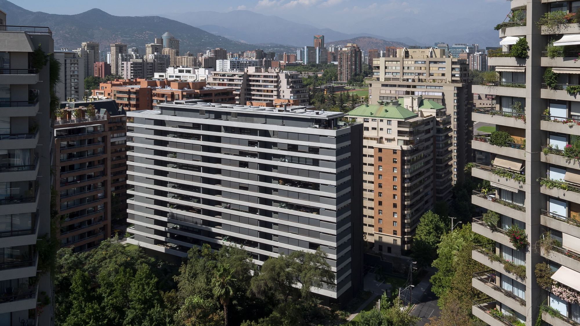 Tower with horizontal grey balcony bands seen among green-roofed neighbours, with mountains rising behind the city under a clear sky