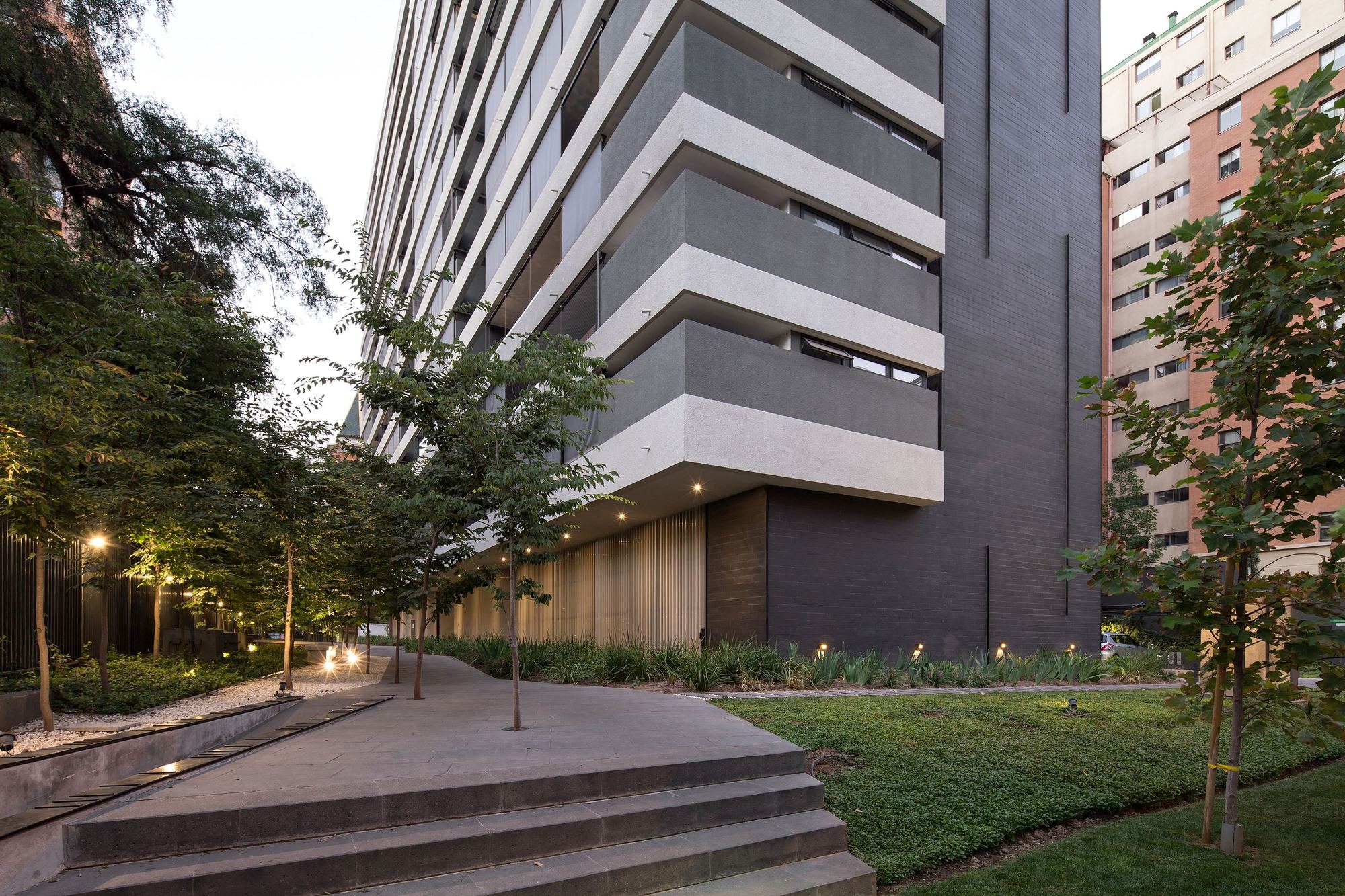 Dusk view of the tower's grey cantilevered balconies above a stepped path, young trees and a lawn, with ground lights glowing