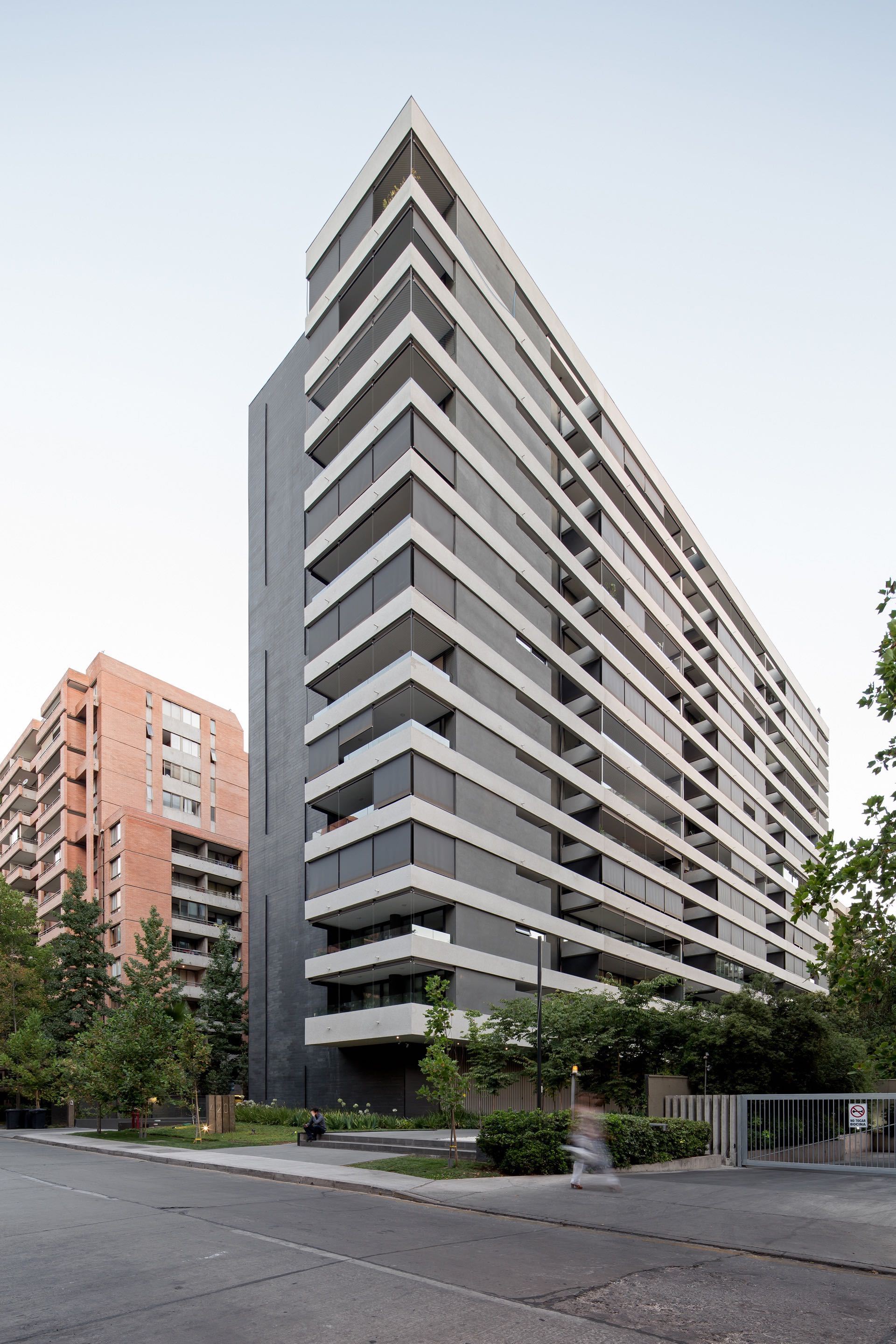 Corner view of the tall dark tower with white stepped balcony slabs, a pedestrian crossing the sidewalk at dusk