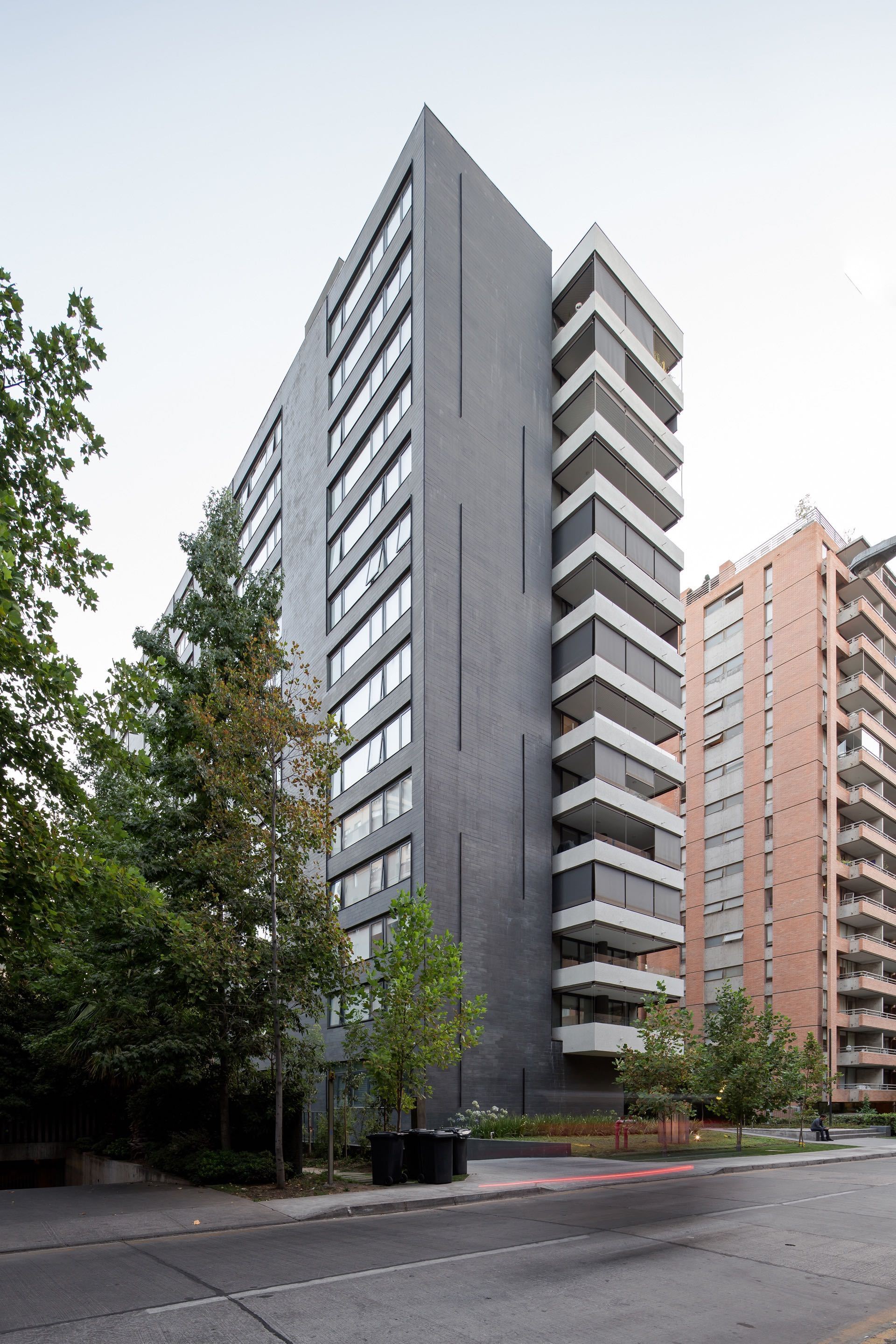 Side elevation of the dark grey tower with narrow vertical slits and stacked balconies beside a brick building, trees in the foreground