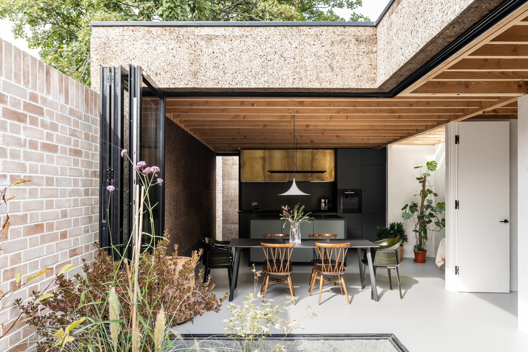 Open dining area with folding black glass doors, timber ceiling and cork walls beside a pale brick garden wall and tall grasses