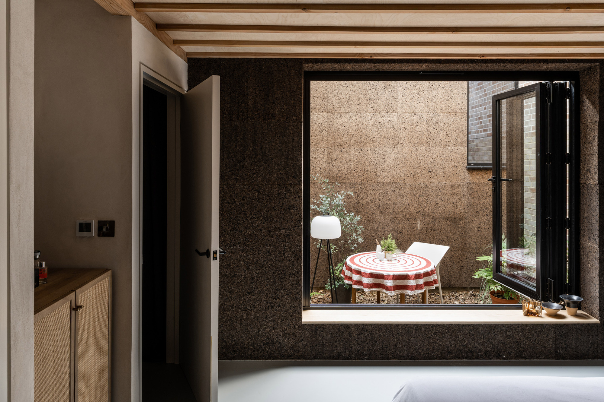 Cork-lined window opening framing a small courtyard table with red striped cloth, with a folding glazed door swung open
