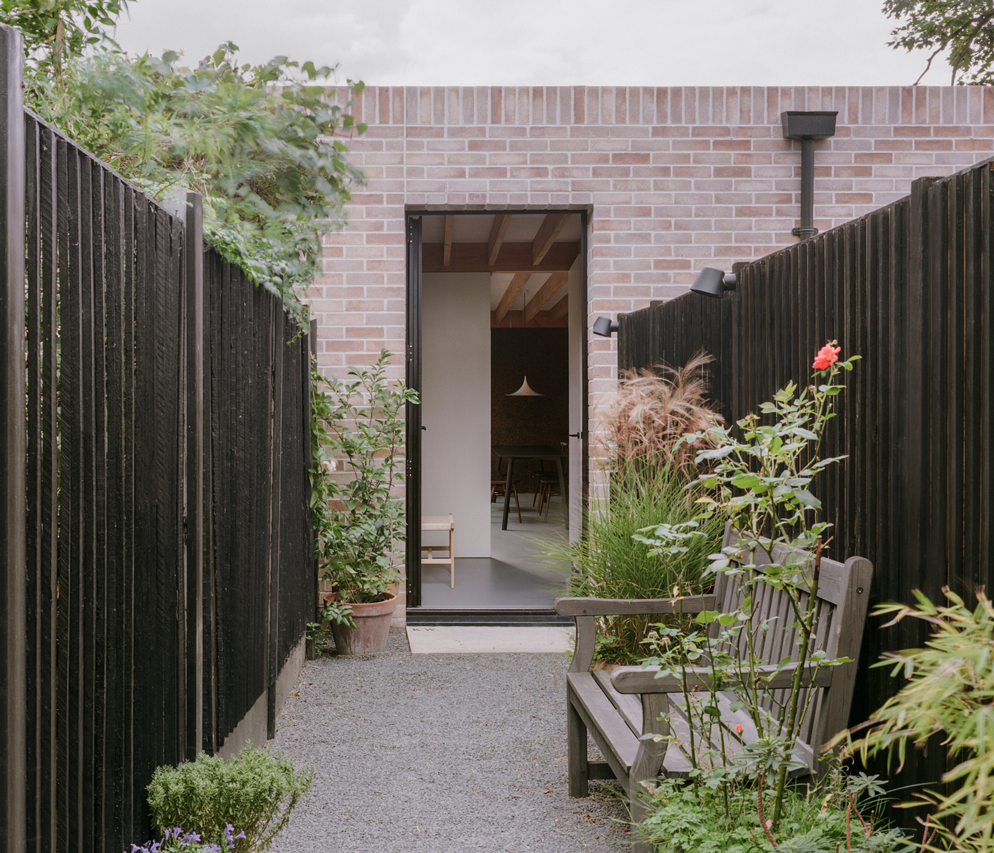 Gravel path between charred timber fences leading to an open door in a pink brick wall, with a wooden bench and roses
