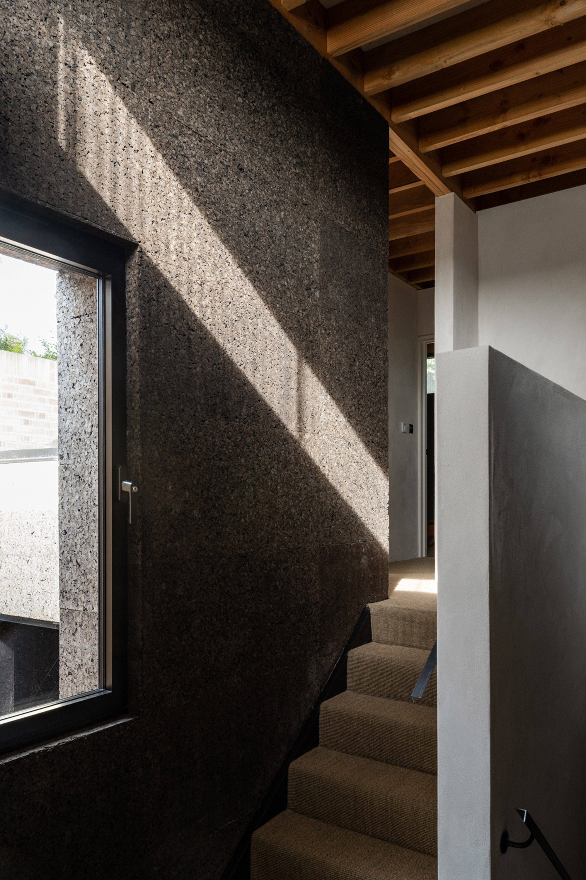 Sunlight cutting diagonally across a dark cork stair wall with a small window, beside carpeted steps and exposed timber joists