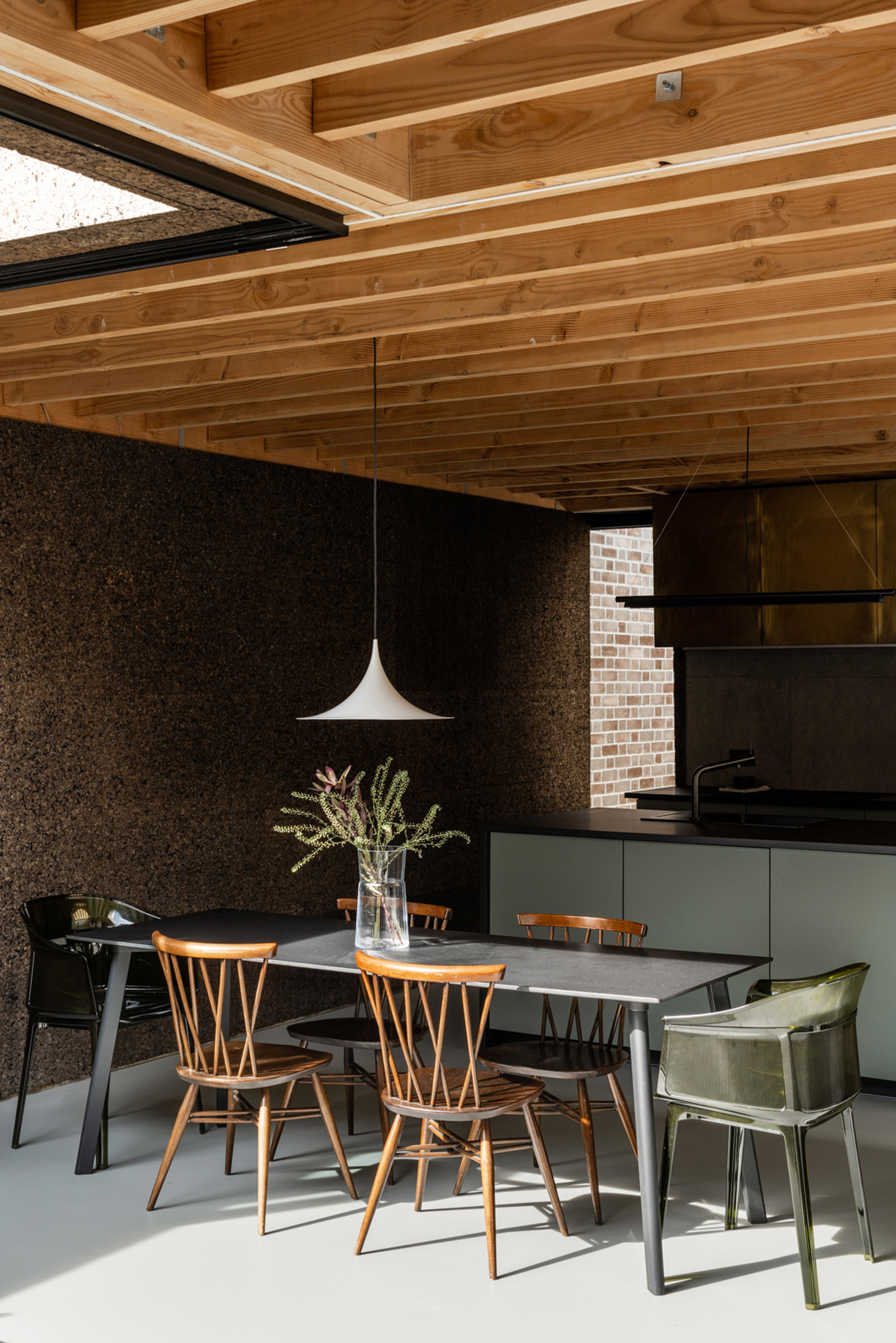 Dining table with spindle-back chairs under a white pendant lamp, set against a cork wall below exposed timber joists